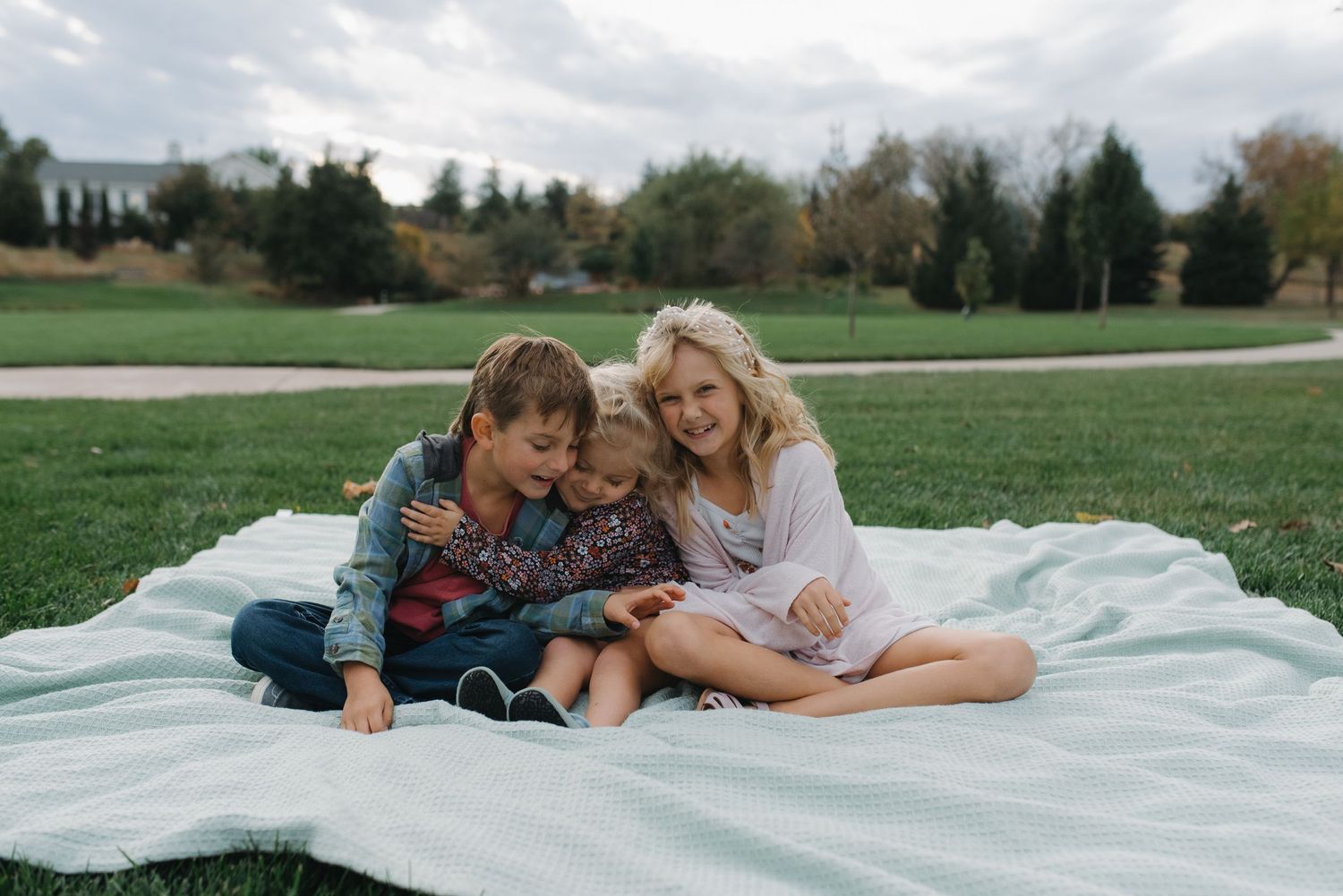 Three children sitting together on a blanket in a green park, smiling and hugging on a cloudy day.