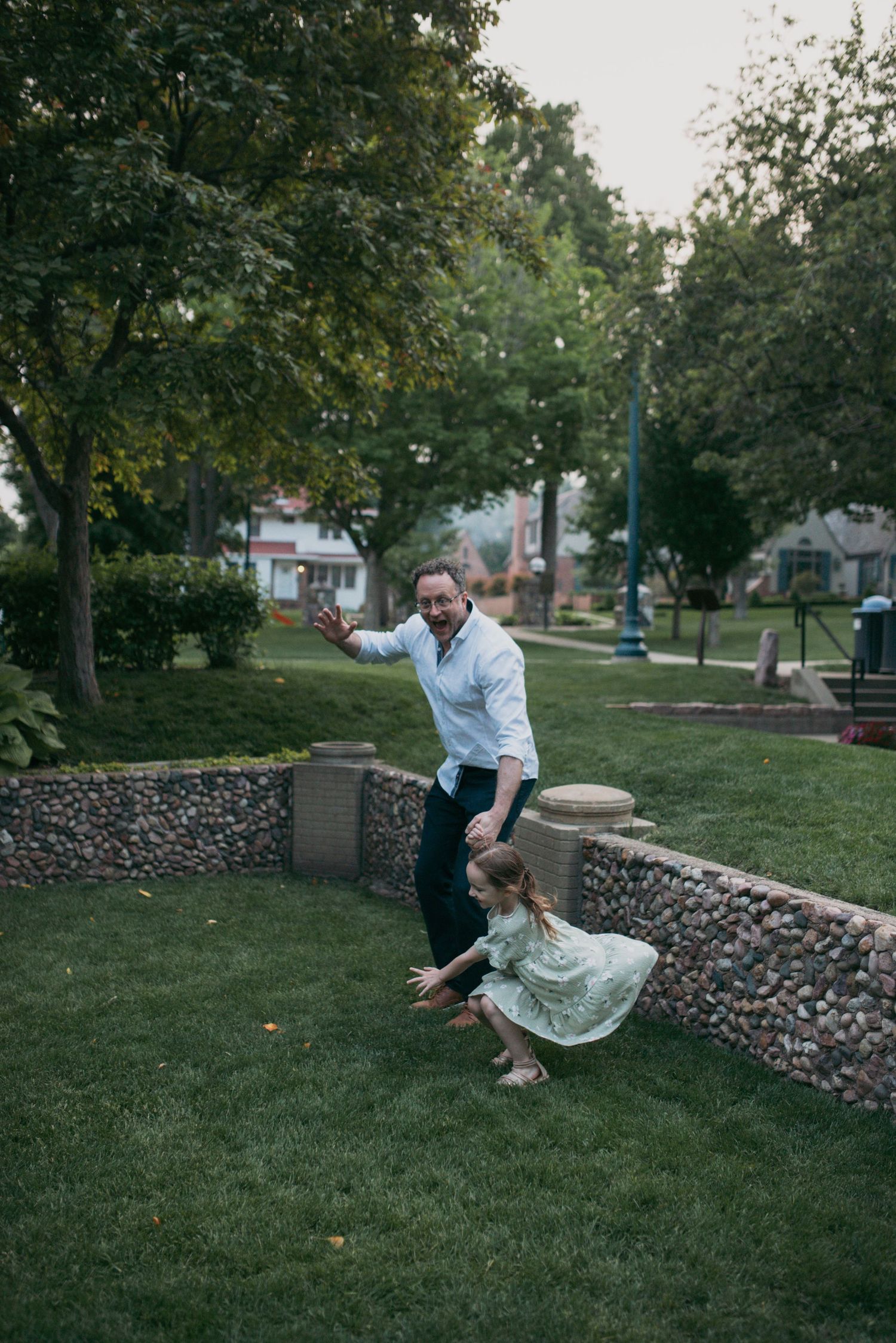 A father and young daughter playing and running together on green grass in a park setting.