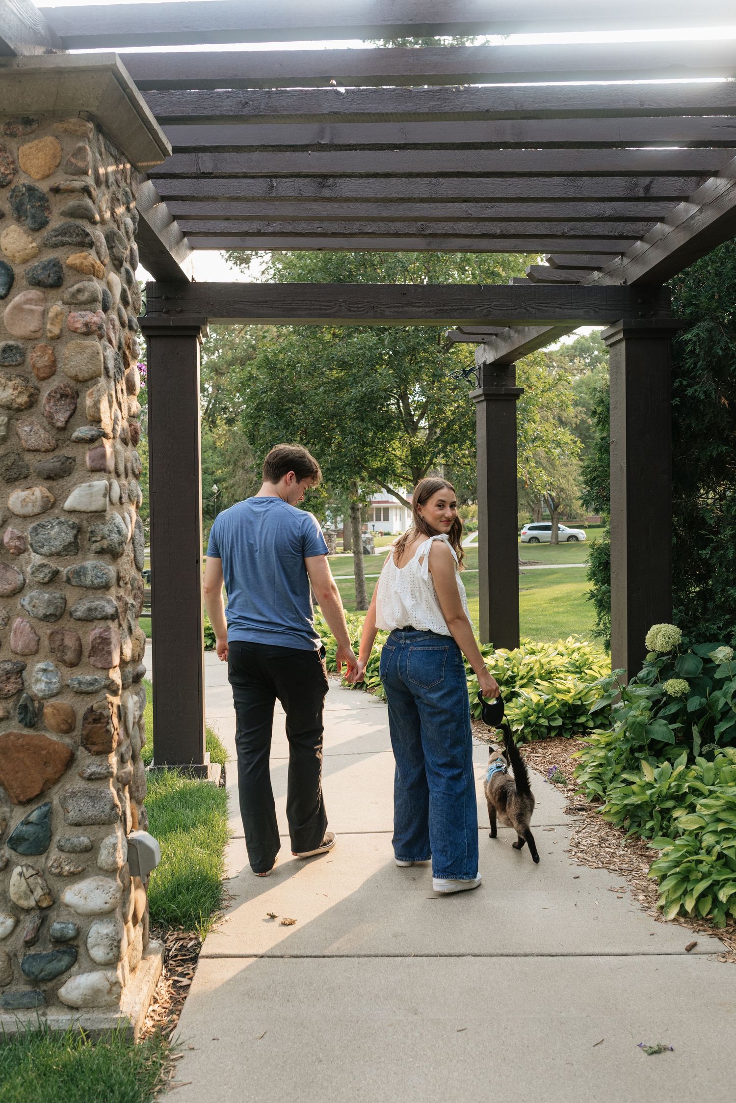 Couple holding hands walking dog under pergola with stone pillars in lush green garden.