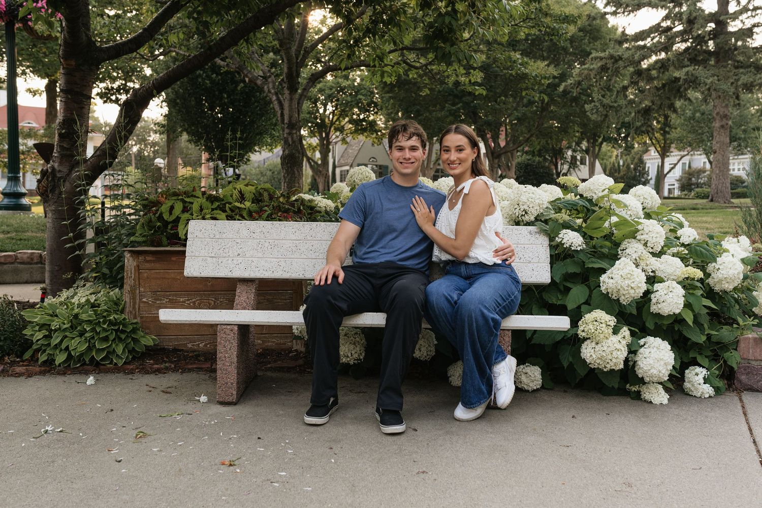 Couple sitting together on a white park bench surrounded by white hydrangea flowers in a lush green park.