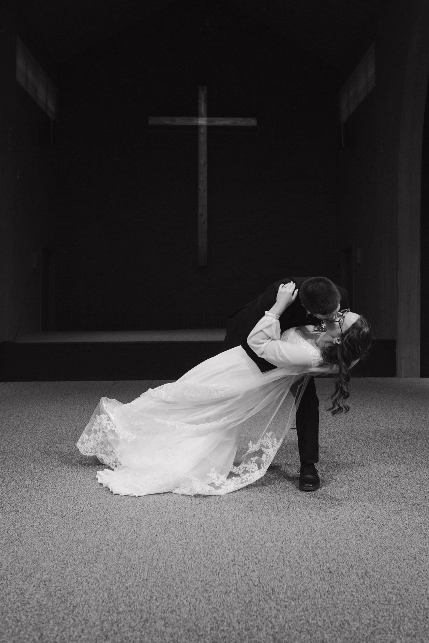 Bride and groom share romantic dip kiss in black and white photo inside church with cross backdrop.