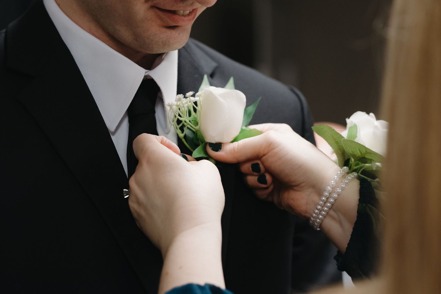 Woman pinning a white rose boutonniere on a groom's black suit lapel at a wedding ceremony.