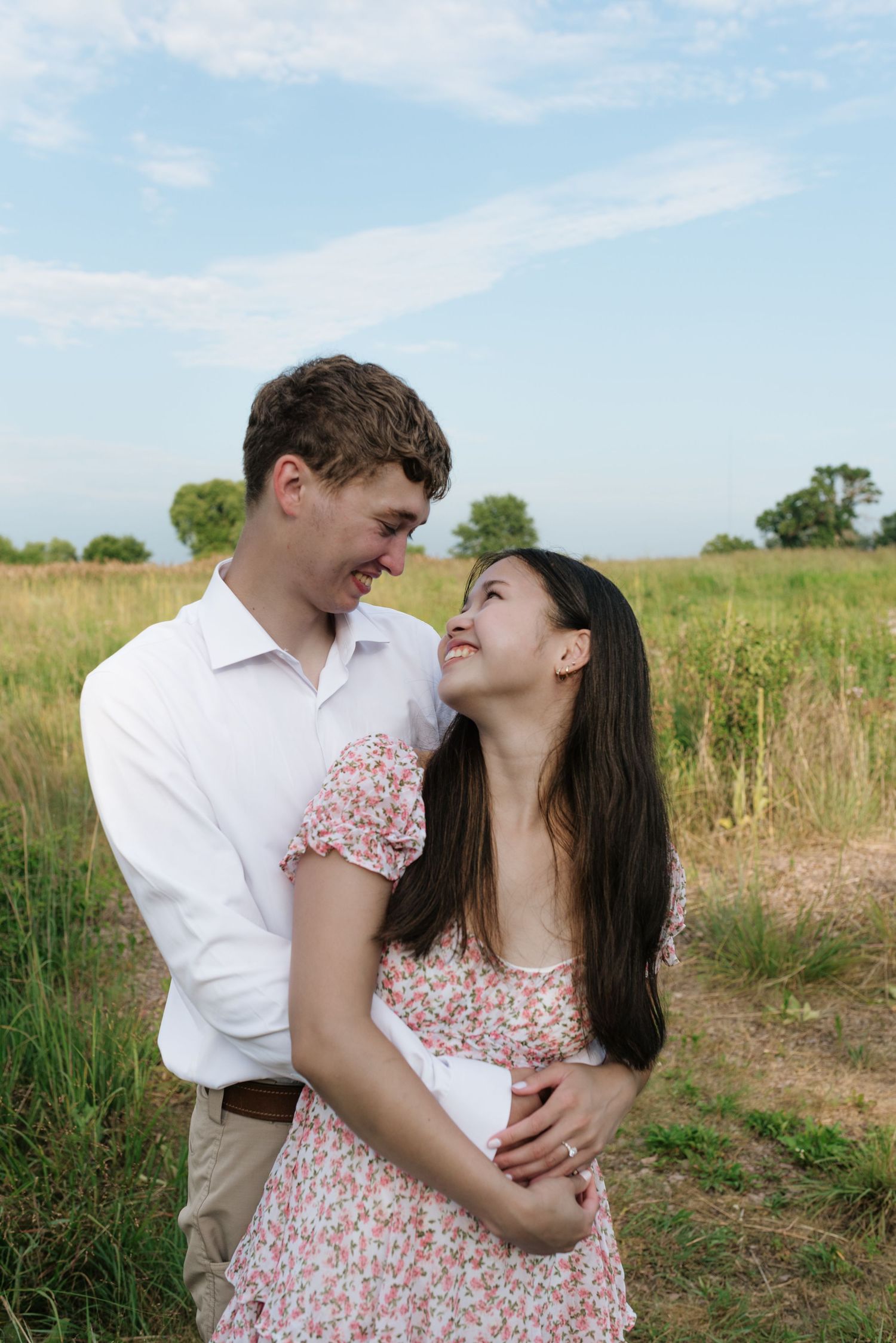 Smiling couple embracing in a sunny green field, woman in floral dress looking up at man.