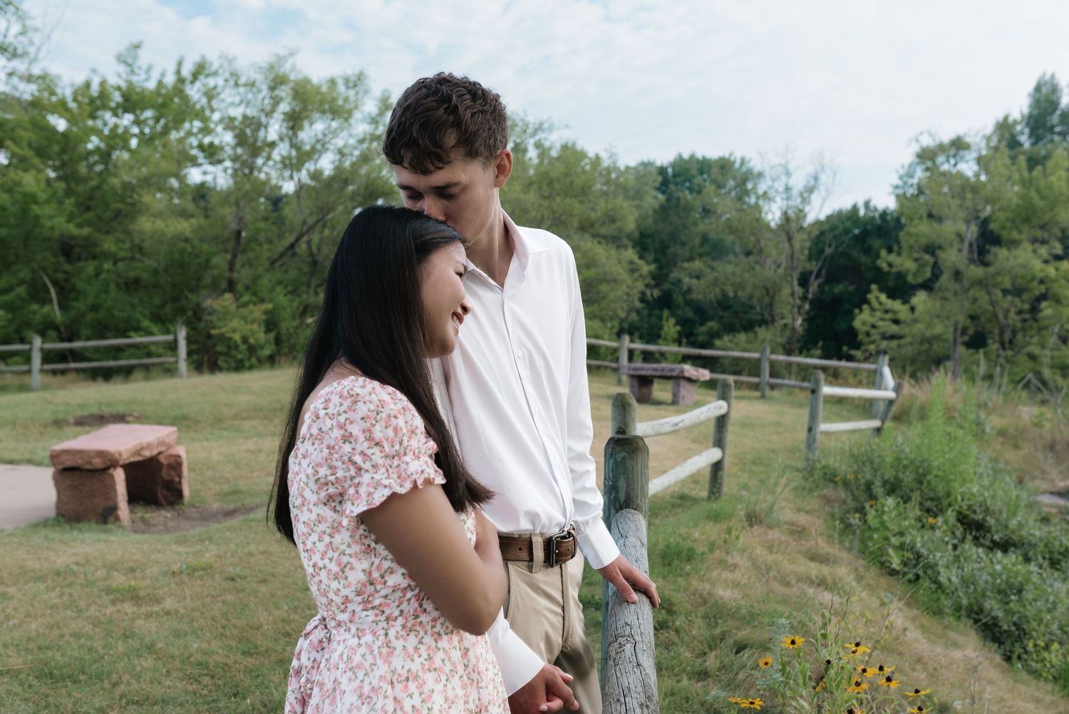 A romantic young couple shares a tender moment by a wooden fence in a lush green park setting.