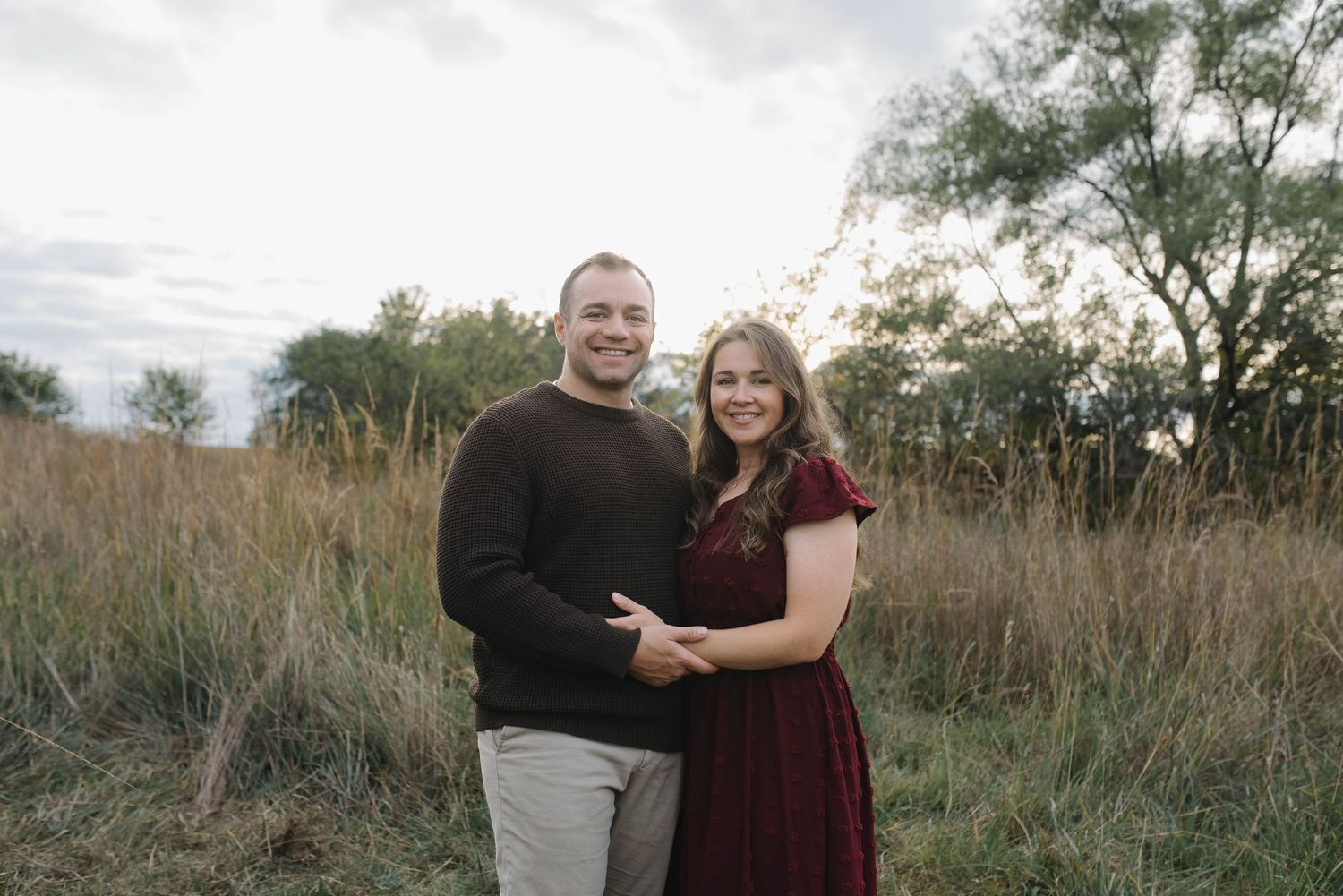 A smiling couple embraces in a golden field, woman in burgundy dress and man in dark sweater.