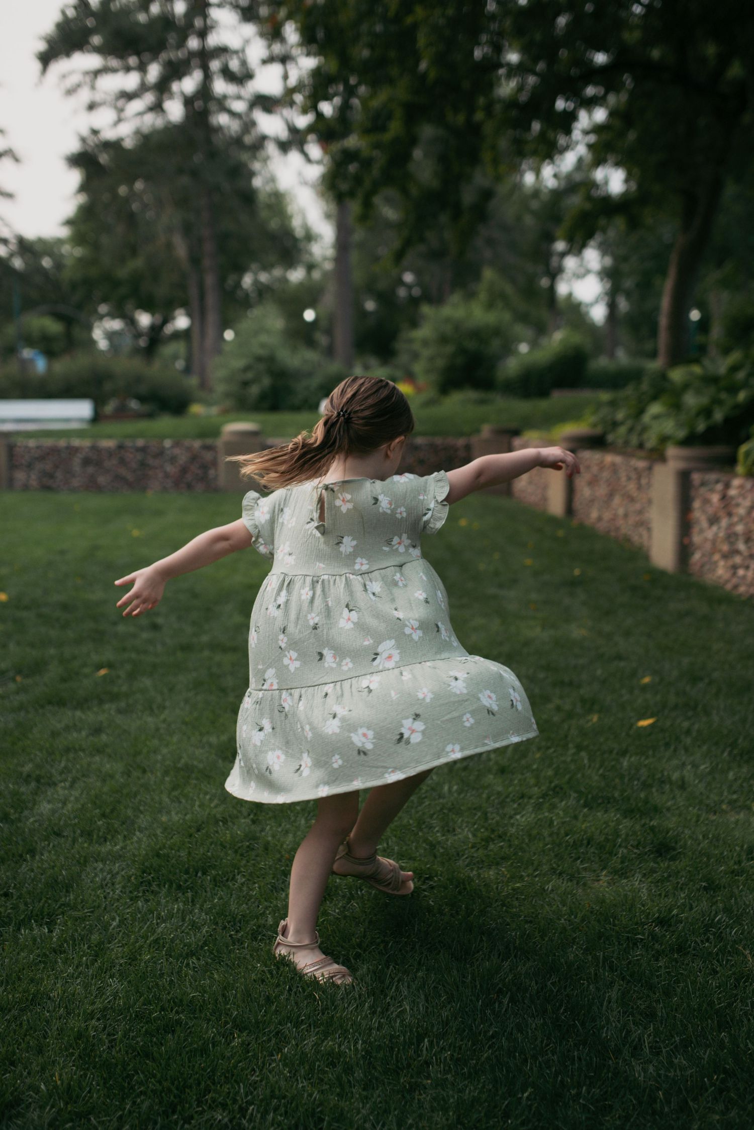 A young girl in a mint floral dress spins joyfully with arms out, her hair flying on a green lawn.