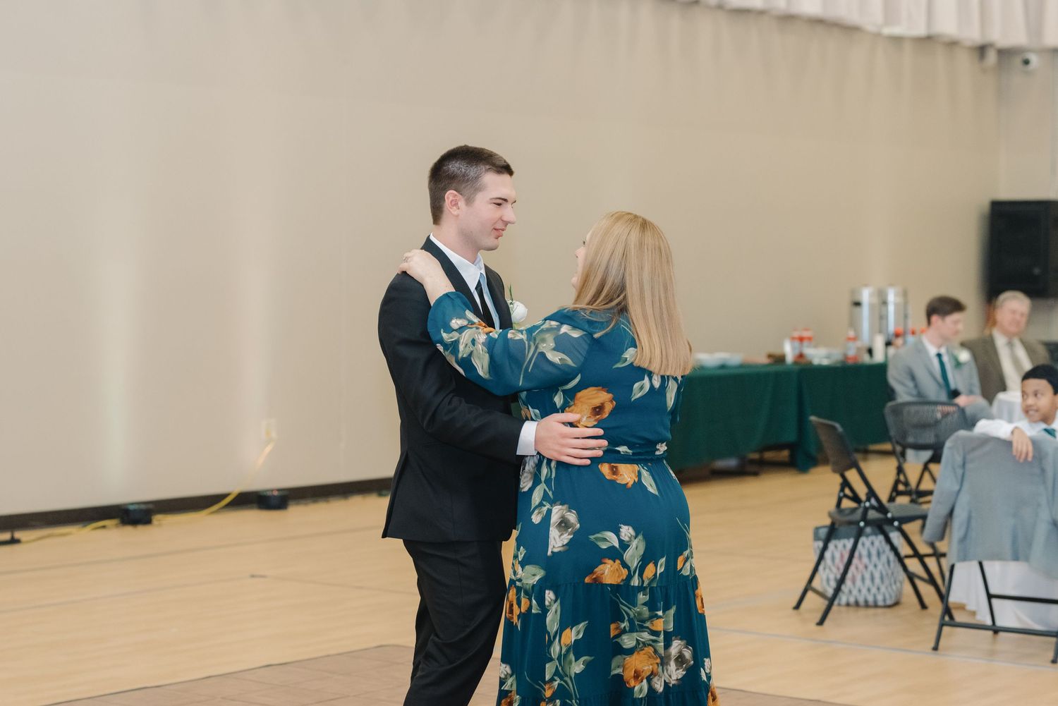Groom in black suit dances with woman in teal floral dress at wedding reception.