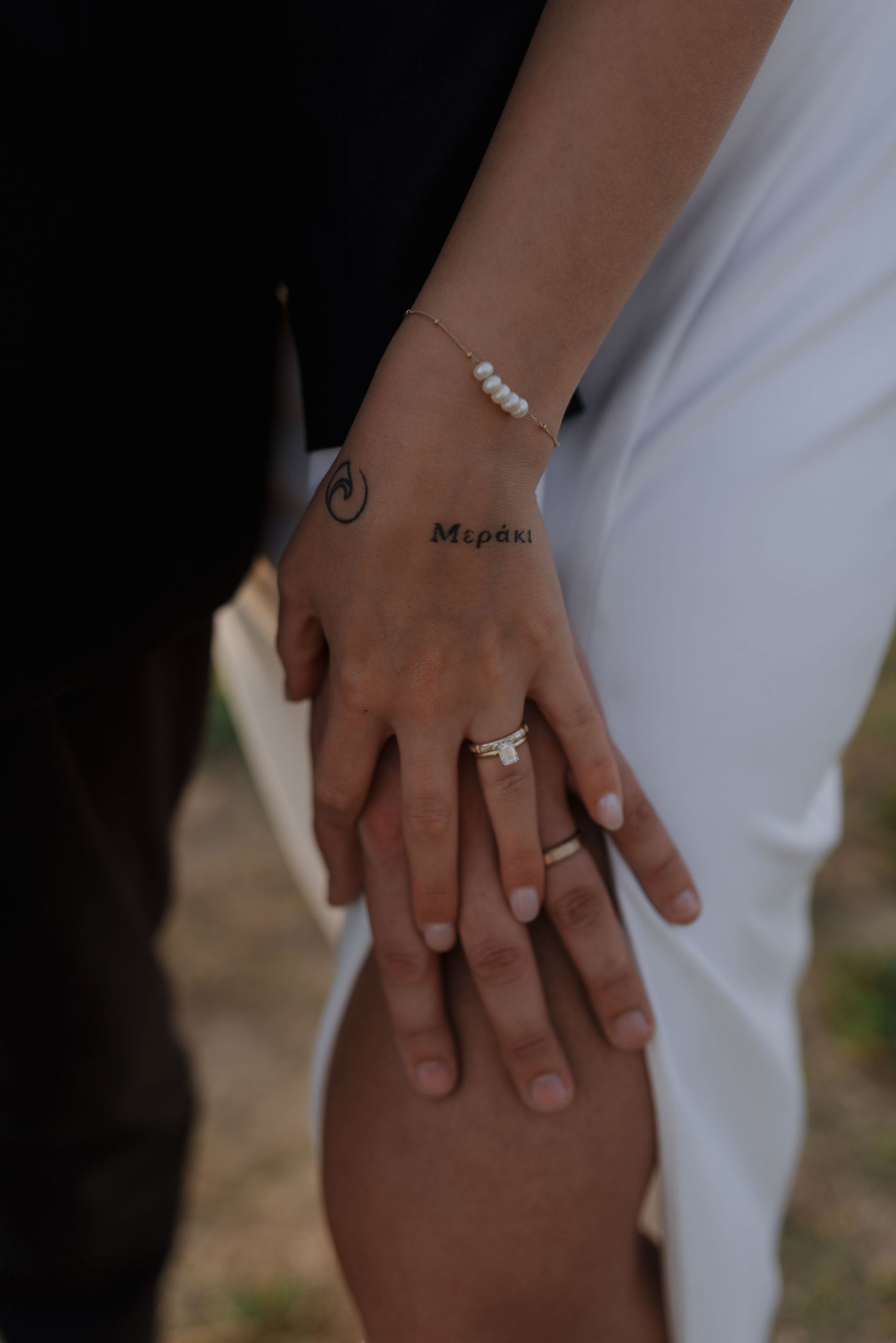 Close-up of bride and grooms hands showing wedding rings, with a Meraki wrist tattoo and pearl bracelet.