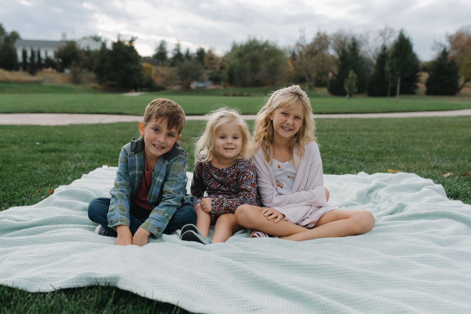 Three young siblings sitting together on mint blanket in green park, smiling happily.