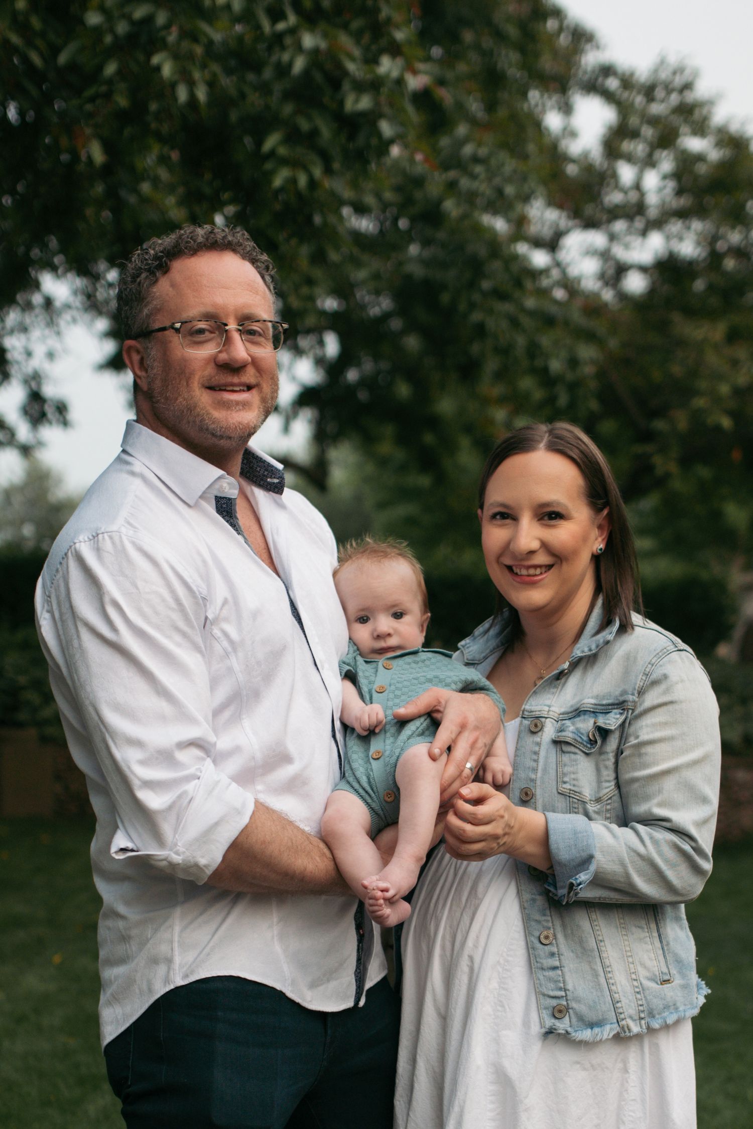 Smiling parents hold baby in teal romper outdoors in green park setting.
