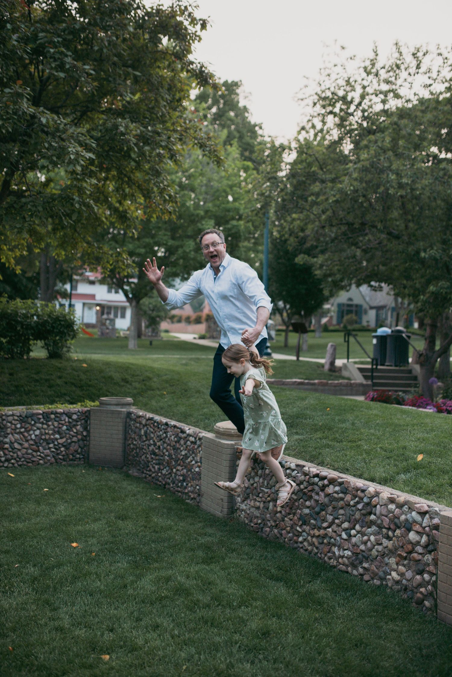 A father and daughter joyfully jumping off a stone garden wall together in a green park.