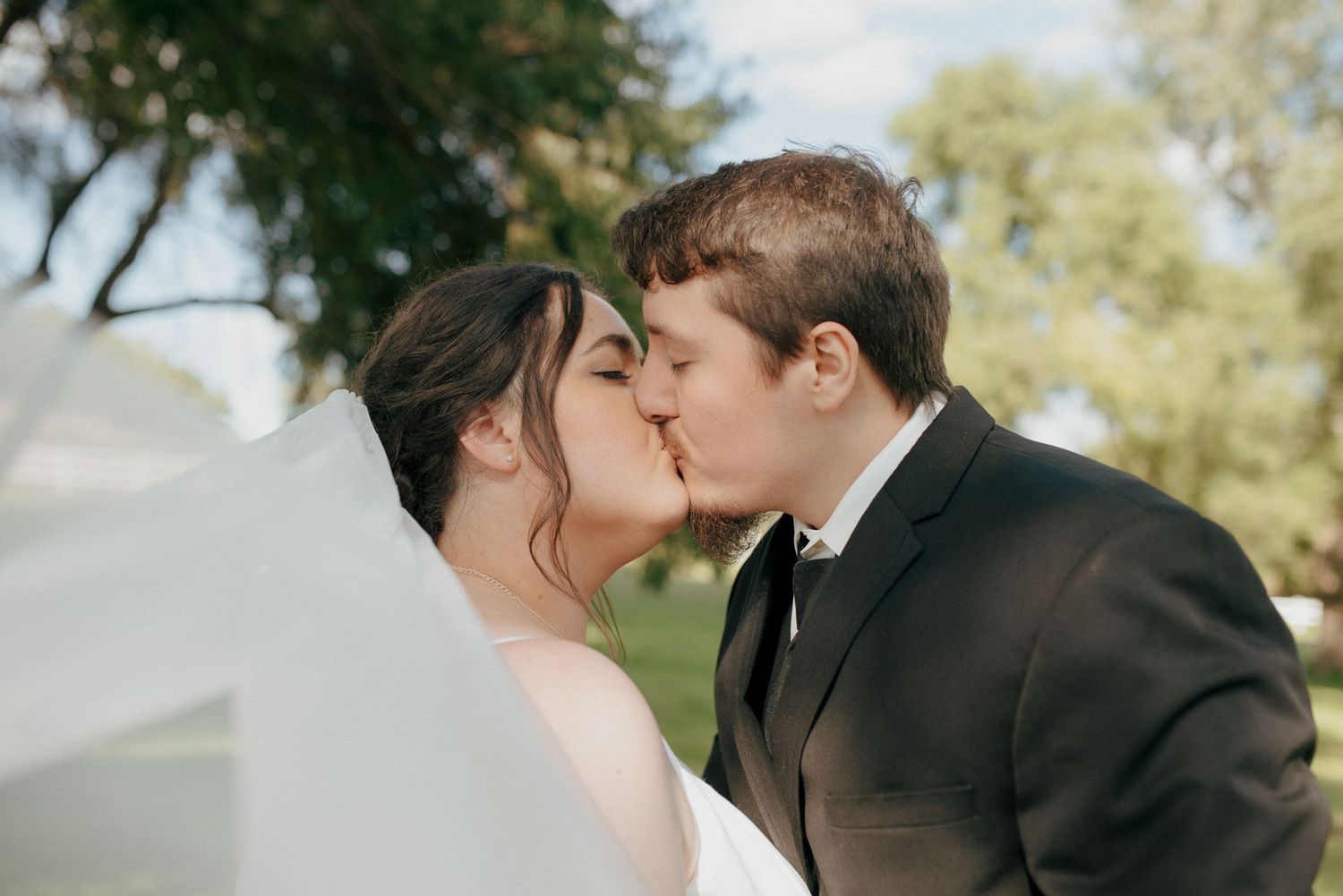A bride and groom share a romantic kiss outdoors with a flowing veil blowing in the wind.