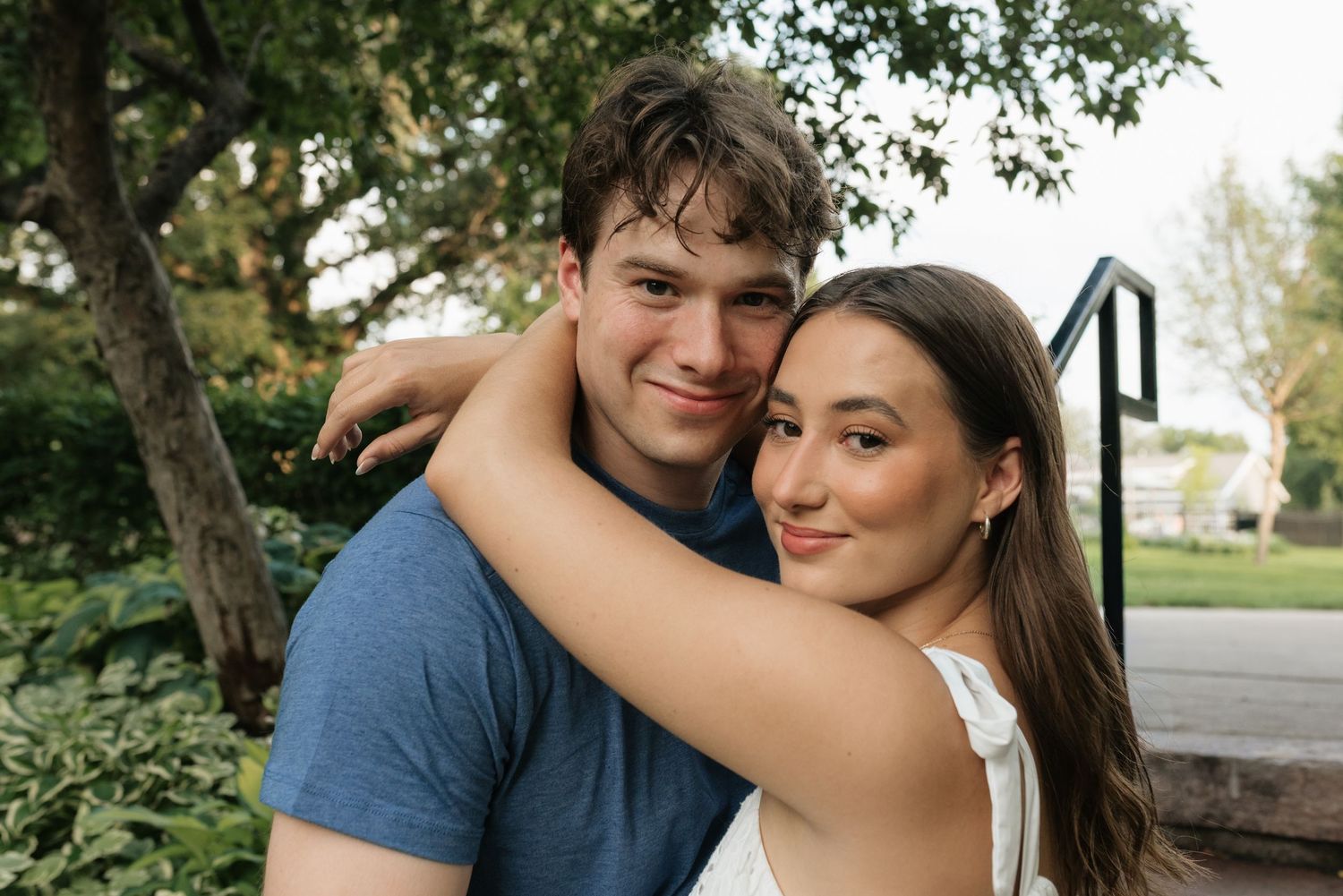 Young couple embracing outdoors, smiling warmly near green trees and steps in a park setting.