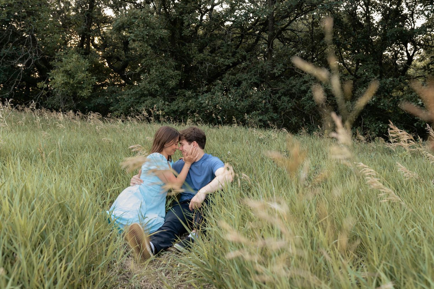 Couple sharing a romantic moment sitting in tall green grass field surrounded by lush trees outdoors.
