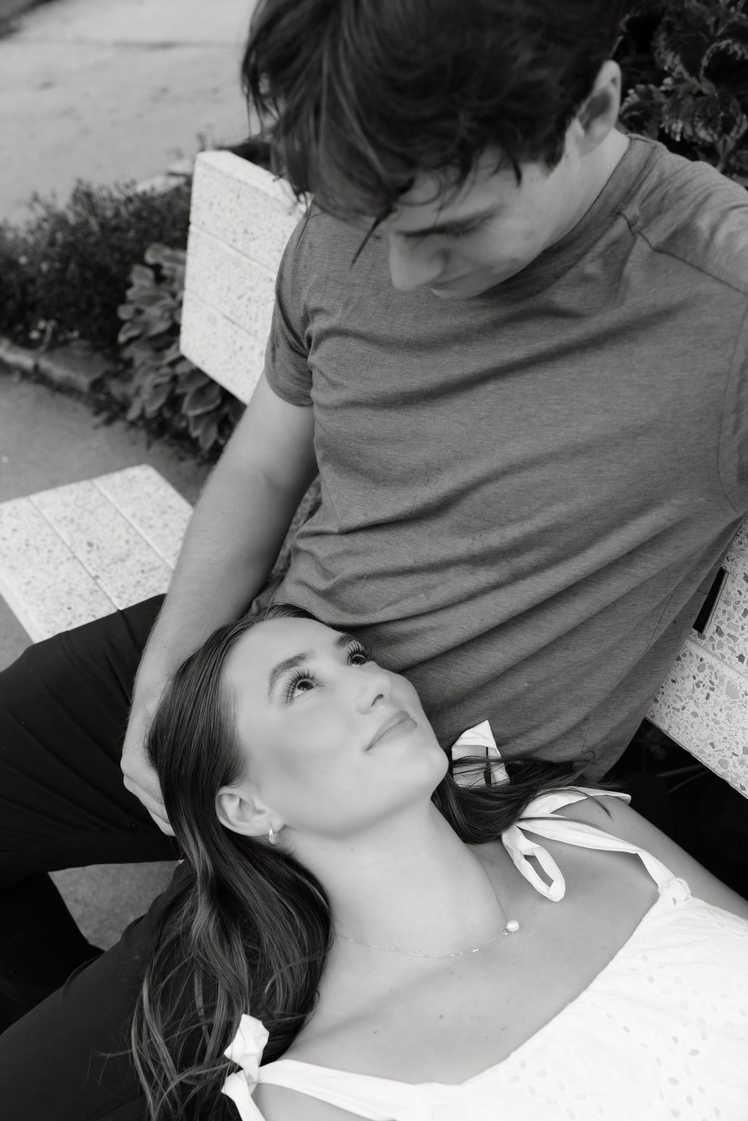 Black and white photo of a woman lying on a bench looking up at a man in a t-shirt gazing down at her.