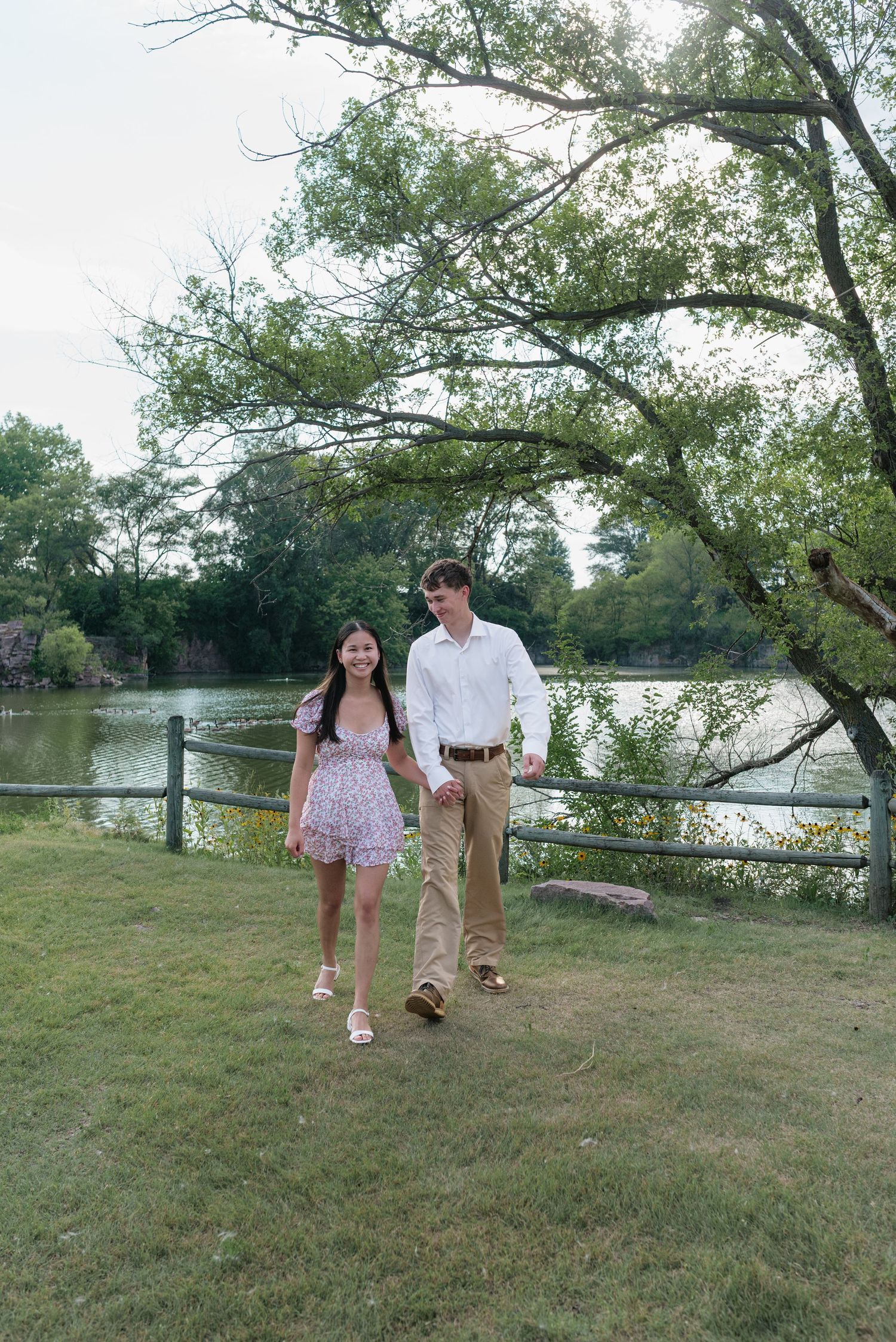 A young couple walks hand-in-hand through a lush park beside a serene lake under large trees.