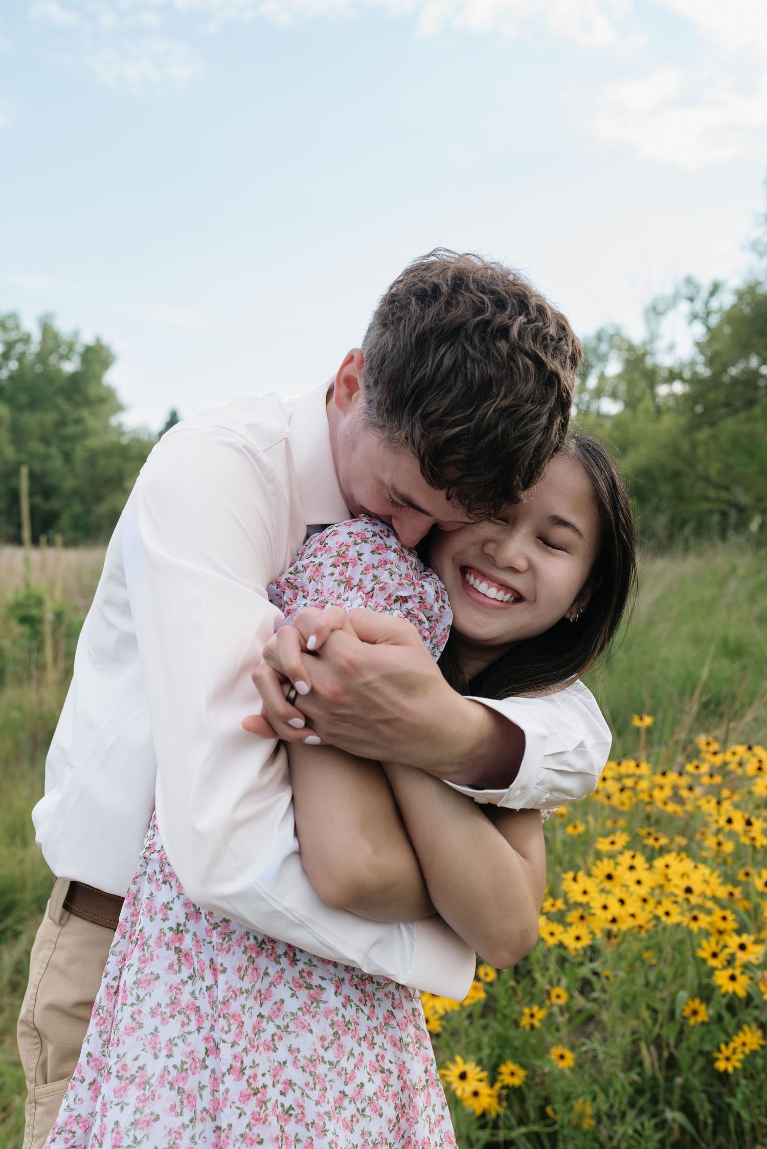 A joyful couple hugging in a field of yellow wildflowers, woman laughing in a floral dress.