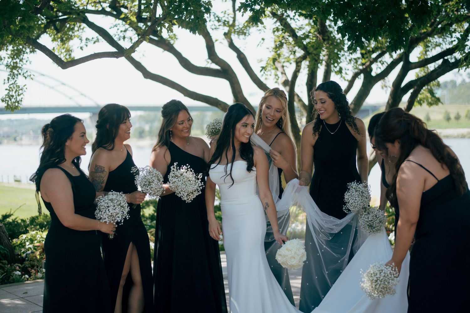 Bride in white gown surrounded by bridesmaids in black dresses holding white floral bouquets outdoors.