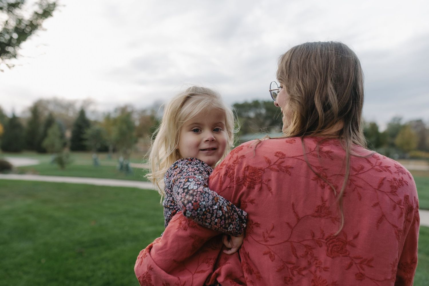 A woman in a red floral top holds a young blonde girl in a park setting.