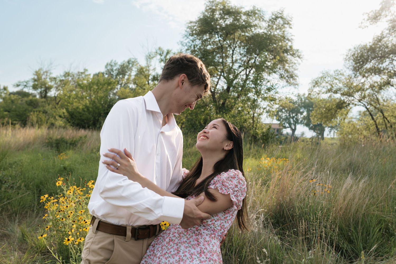Couple laughing and embracing in sunny field with yellow wildflowers and green trees behind them.