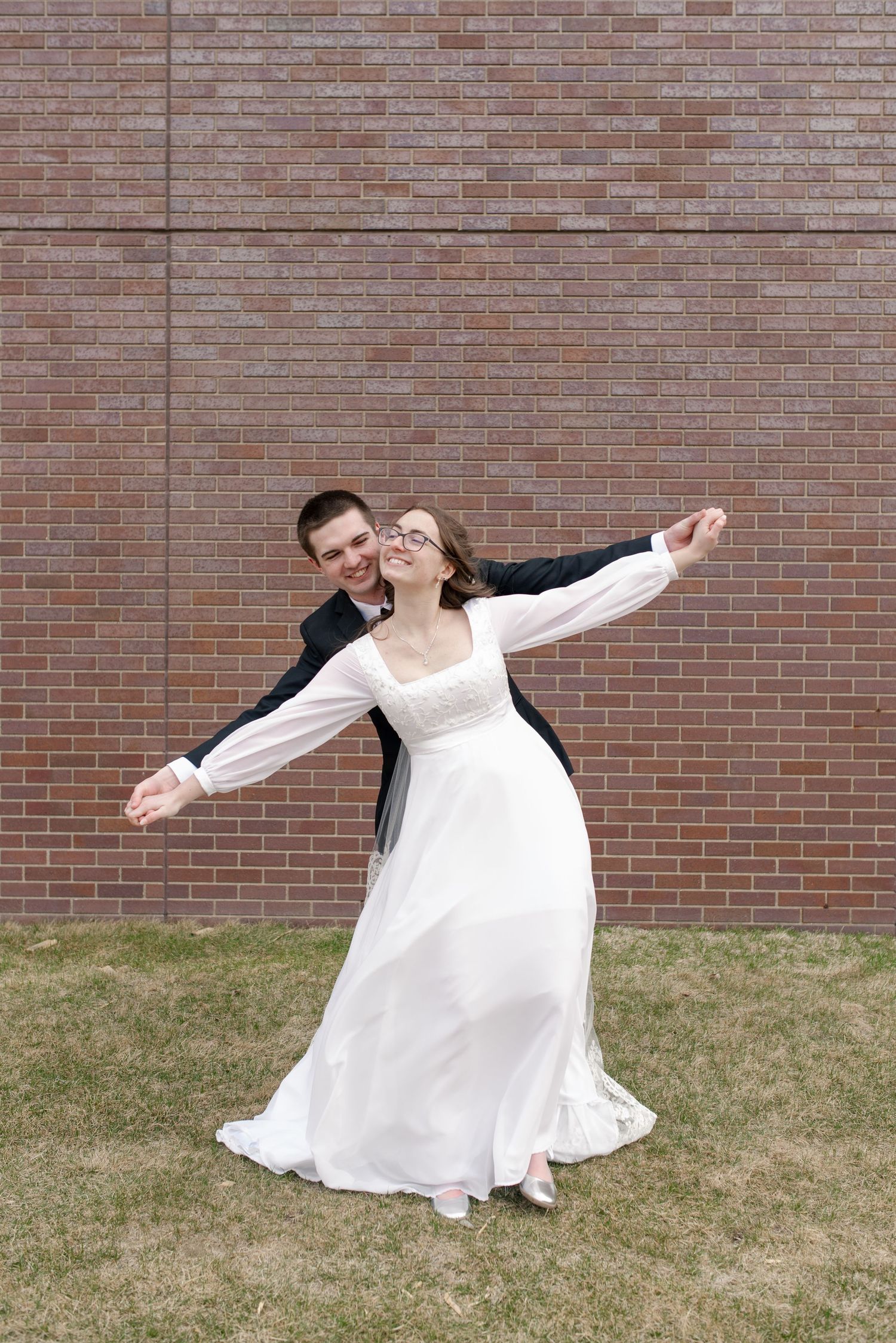 Joyful bride and groom dancing outdoors in front of a brick wall, wearing wedding attire on their special day.