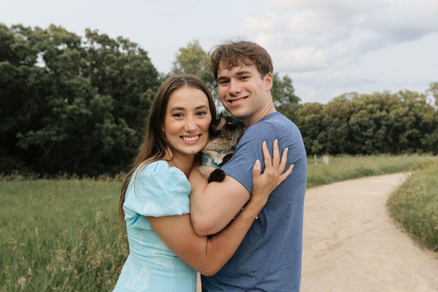 A smiling young couple embraces outdoors on a scenic path surrounded by green fields and trees.