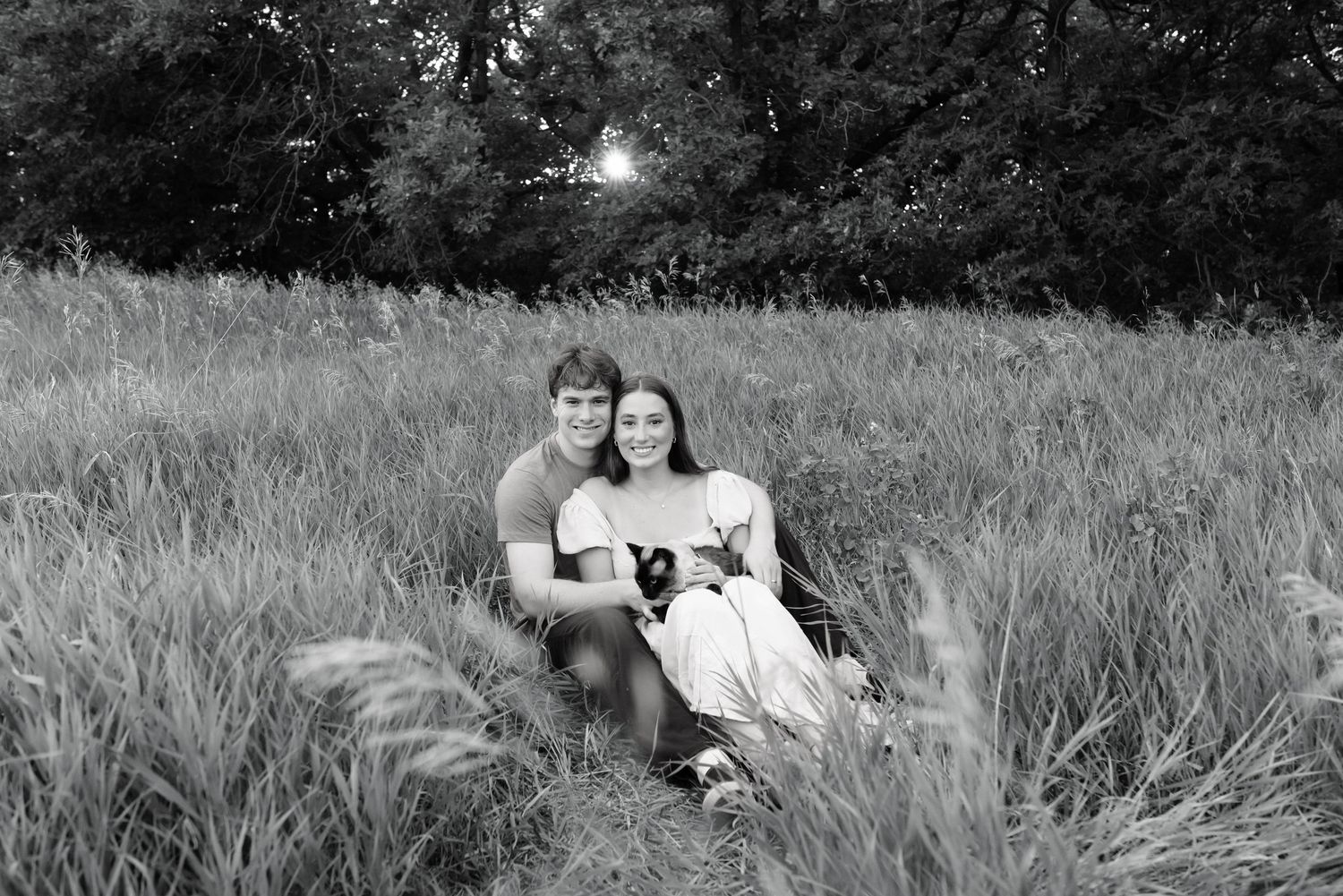 Black and white photo of a couple sitting together in a grassy field, smiling and holding a small dog.