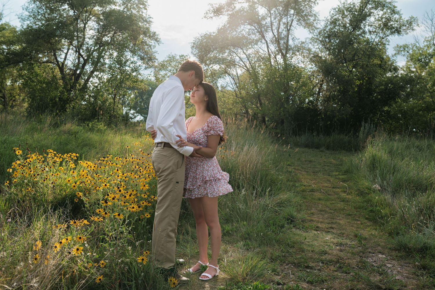 A romantic couple nearly kissing on a nature trail surrounded by golden wildflowers and tall green trees.