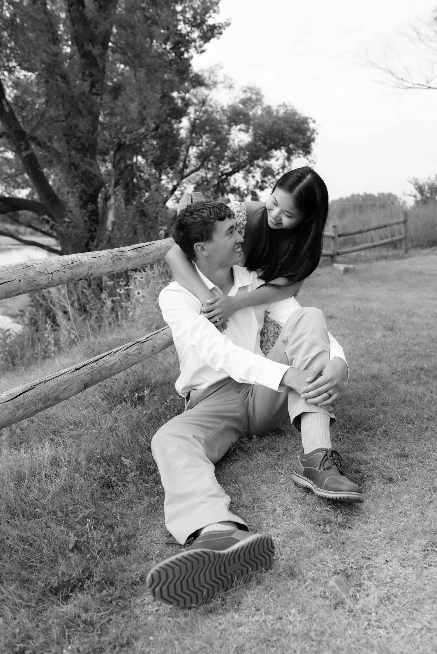 Black and white photo of a couple sitting against a wooden fence, sharing a tender moment outdoors.