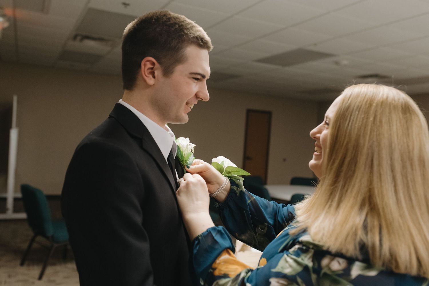 A woman pins a boutonniere on a young man in a black suit before a wedding ceremony.