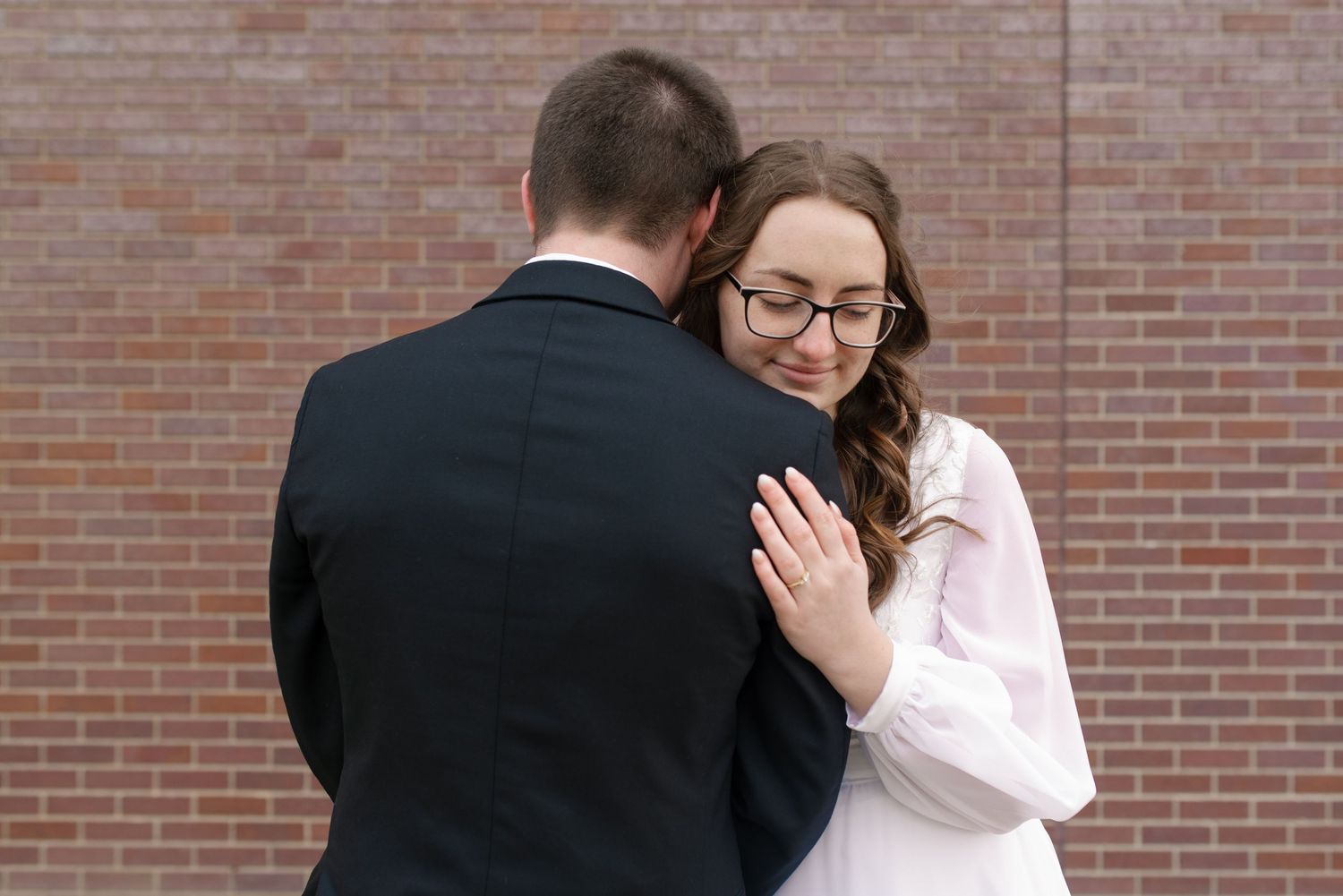 Bride wearing glasses tenderly embraces groom in black suit against a red brick wall at their wedding.