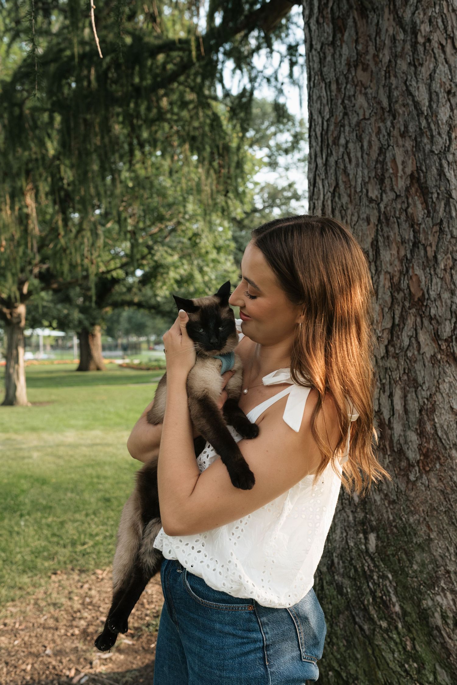 Young woman in white top holding a Siamese cat against a tree in a sunny park.