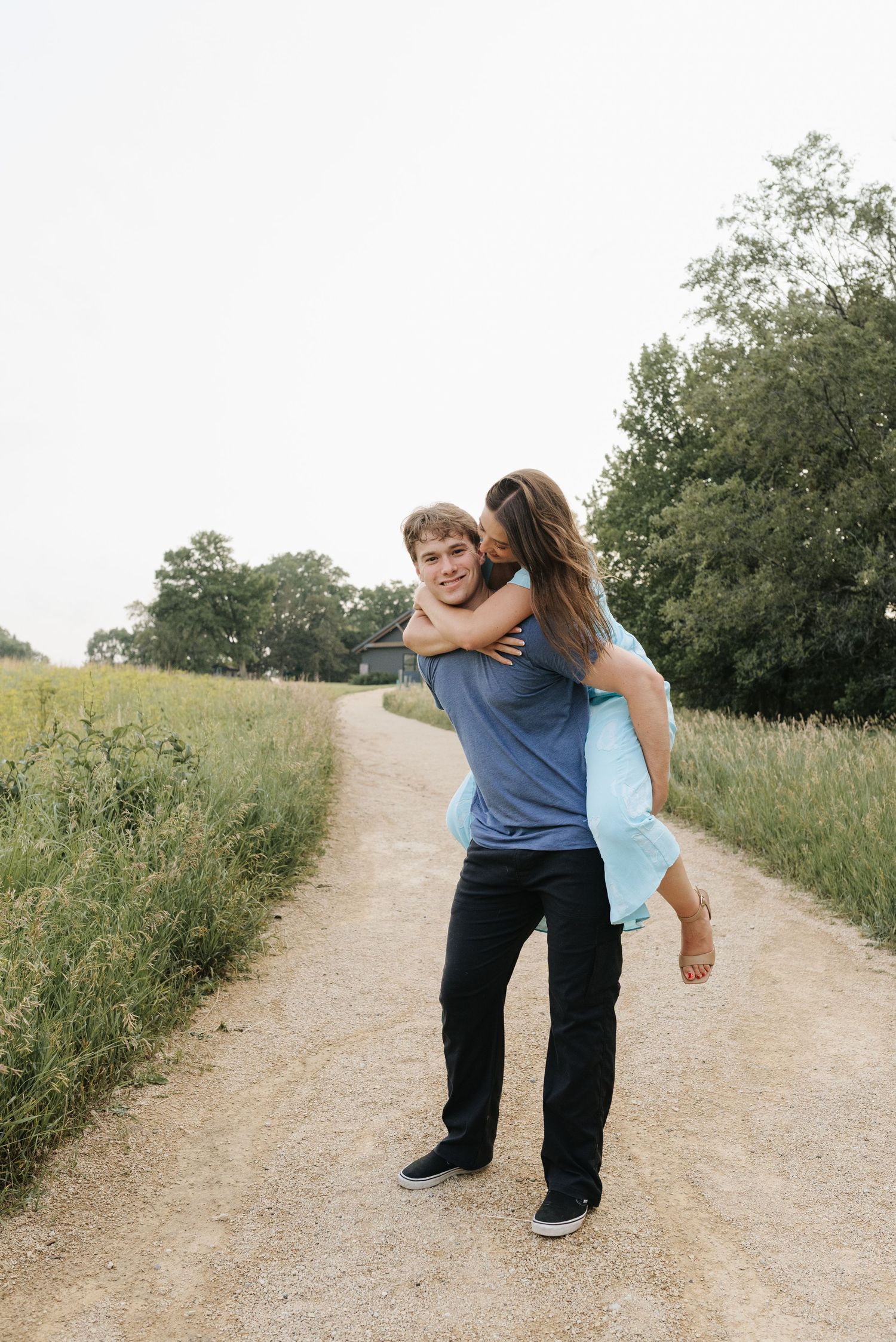 Young couple sharing a piggyback ride on a scenic dirt path surrounded by green fields and trees.