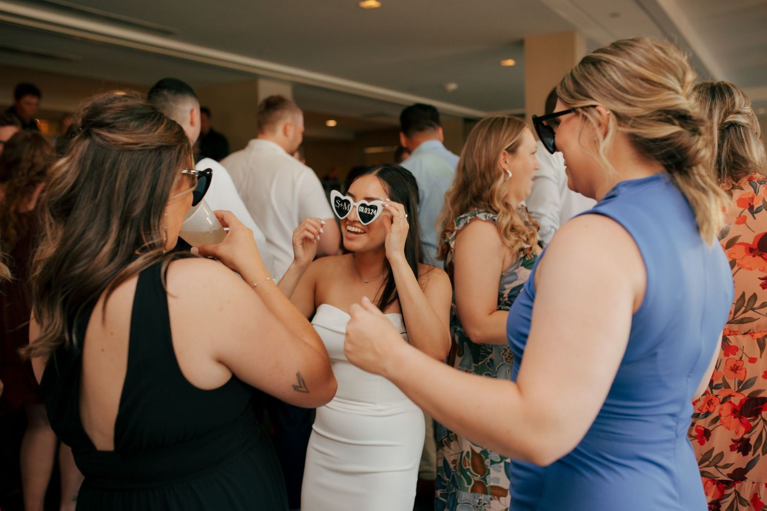 Bride in white dress laughs with friends wearing novelty sunglasses during wedding reception celebration.