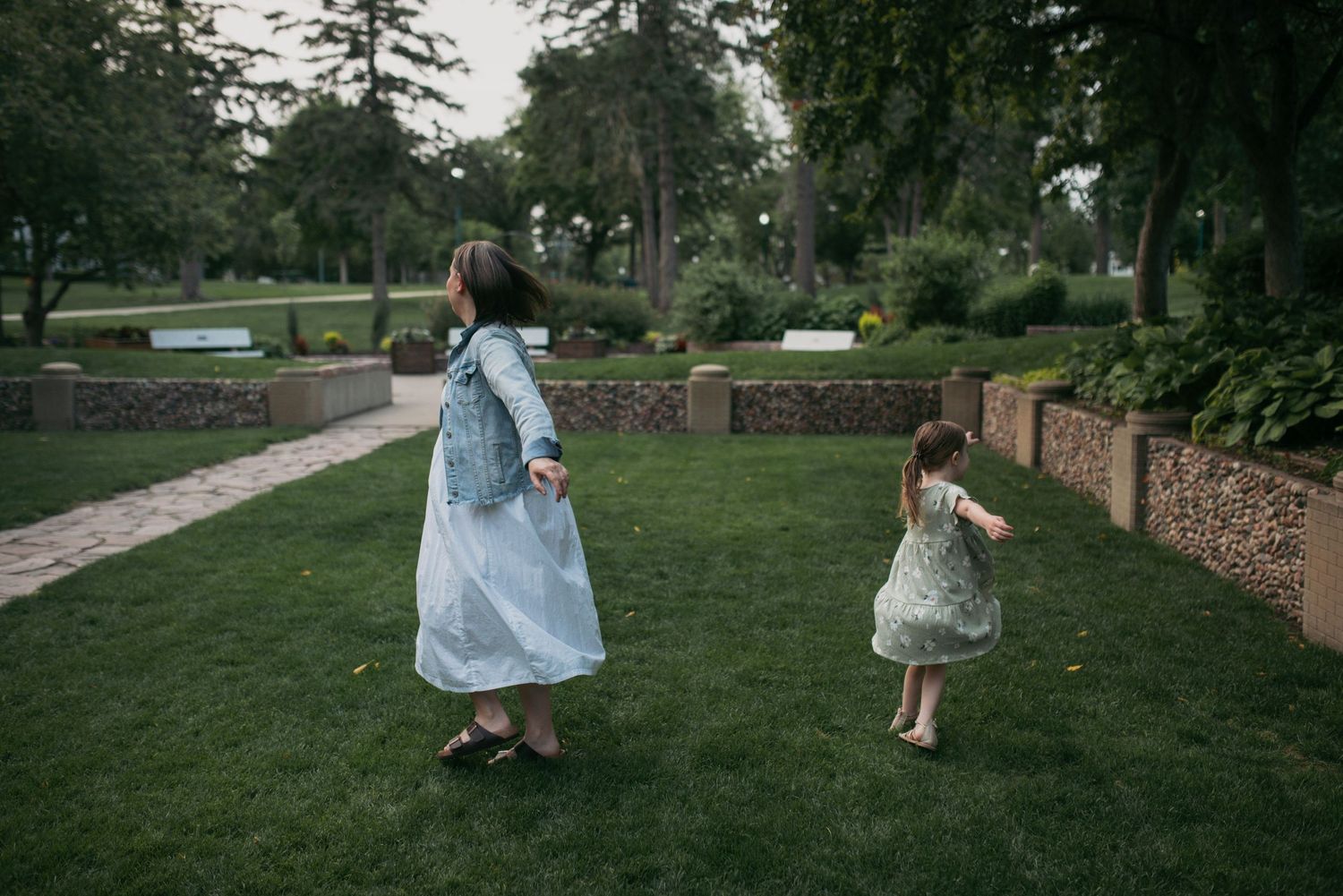 A woman in a white dress and denim jacket chases a young girl in a floral dress across green park grass.