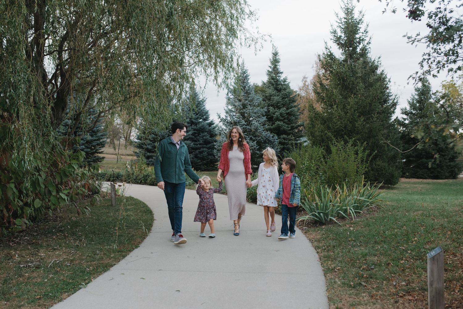 Family of five walking together on a park path surrounded by lush green trees.