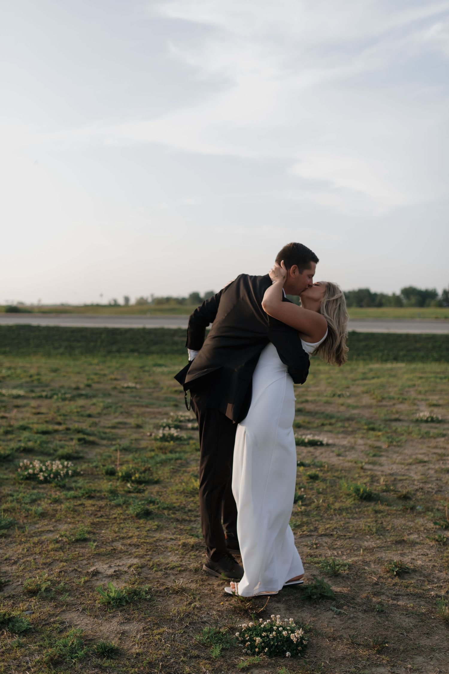 Bride and groom sharing a romantic kiss outdoors in an open field at sunset during their wedding day.