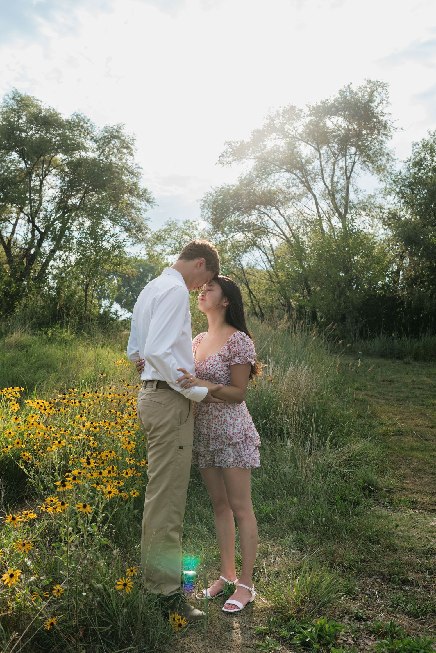 A couple shares a tender moment on a sunlit path beside vibrant yellow wildflowers in a green meadow.