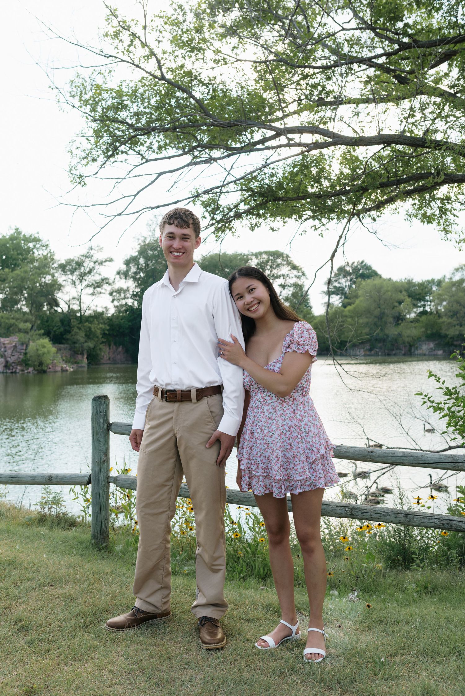 Young couple posing by a wooden fence near a pond, man in white shirt and khakis, woman in floral dress.