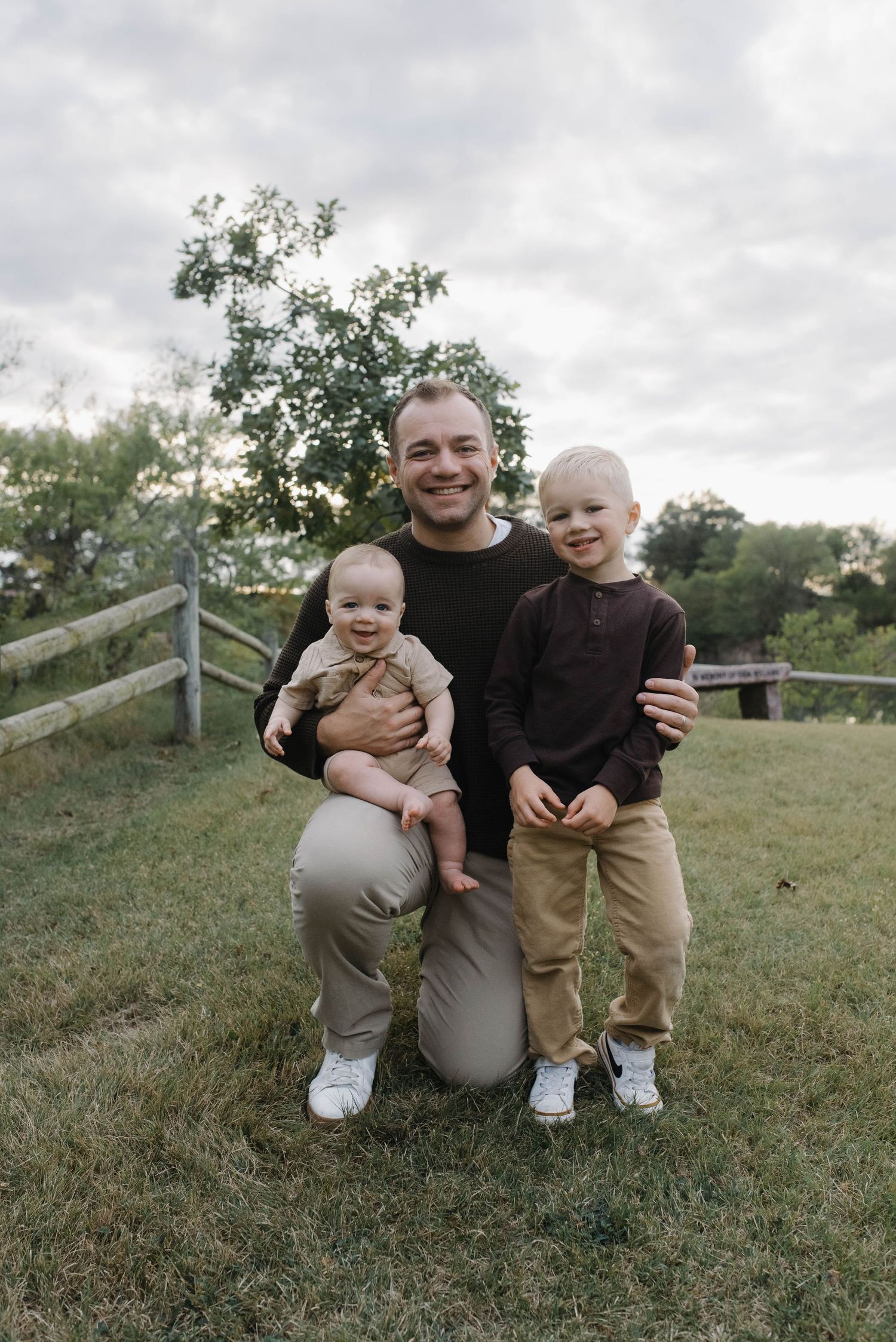 Smiling father crouching on green grass holding infant while young toddler boy stands beside him.