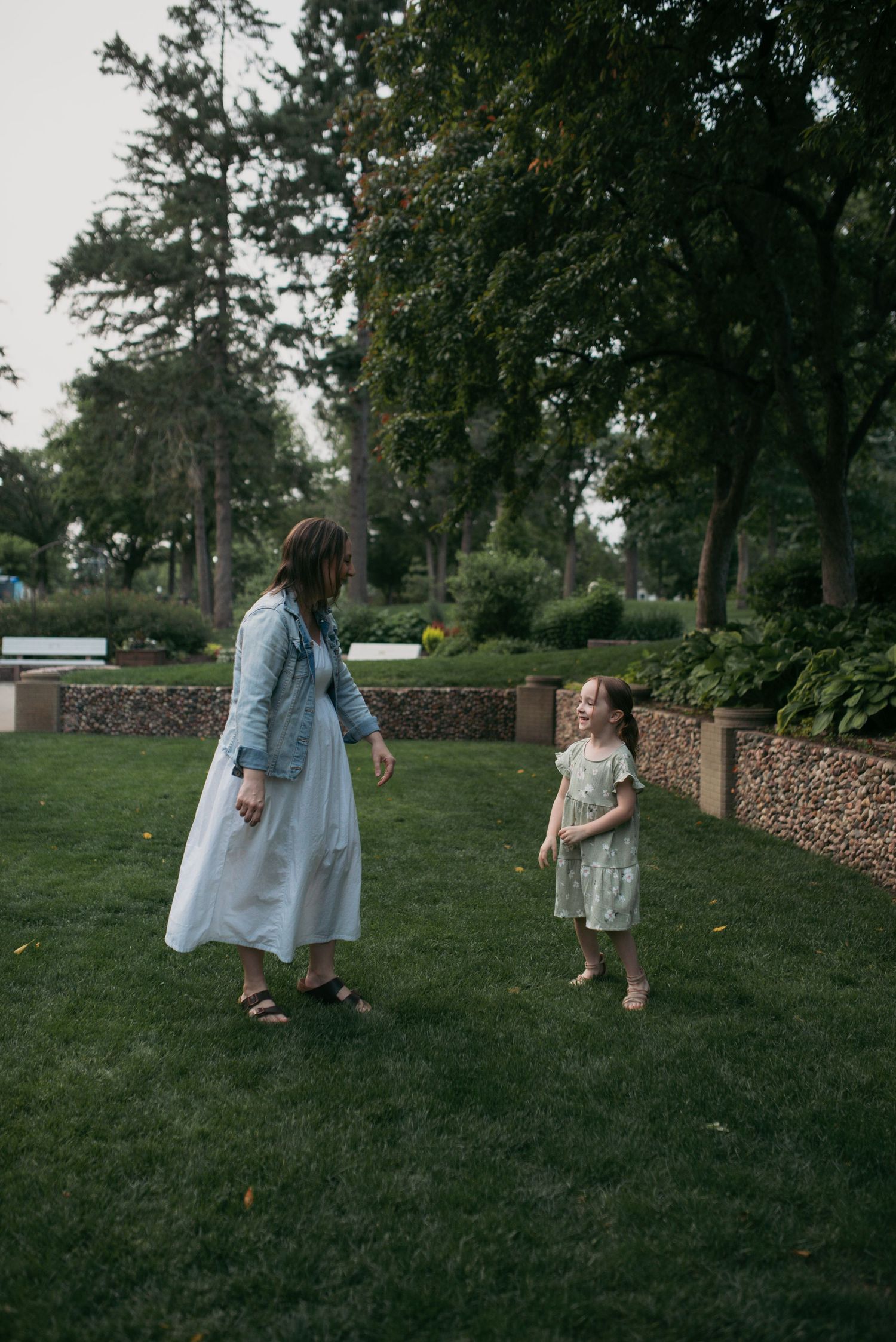 A mother in a white maxi dress and denim jacket faces her young daughter in a floral dress in a green park.