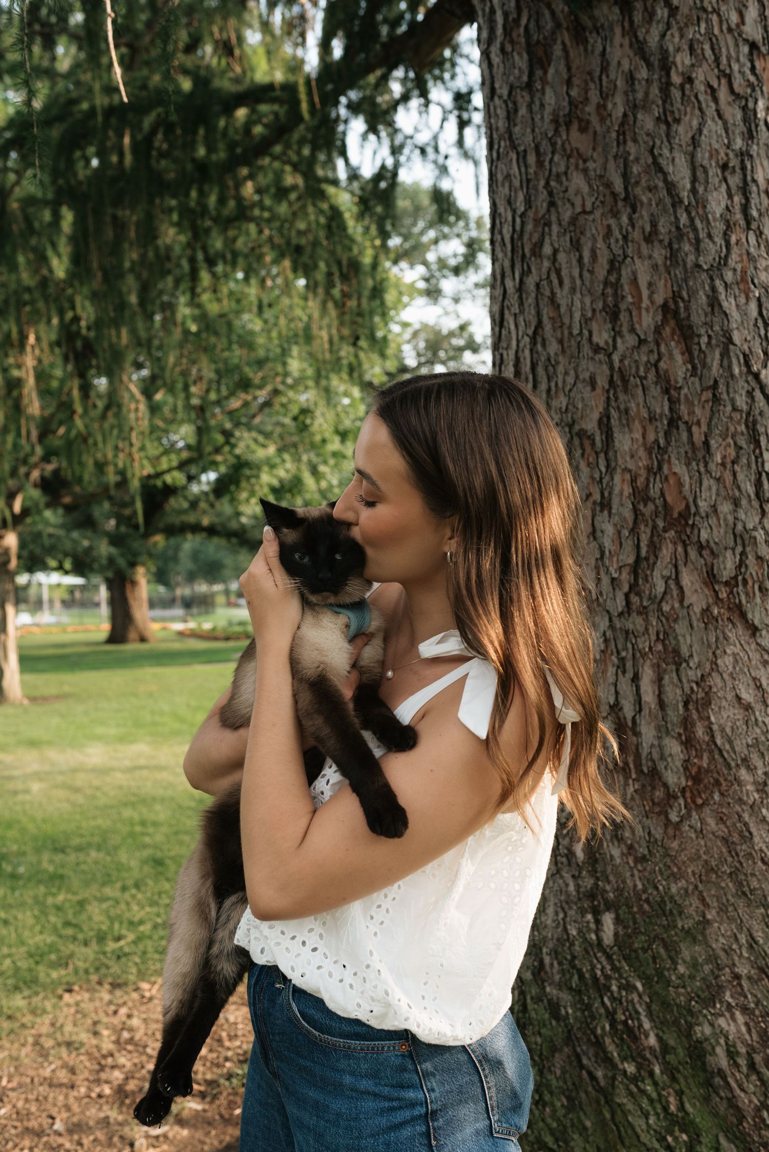 A young woman in a white top kisses her Siamese cat while leaning against a tree in a lush green park.
