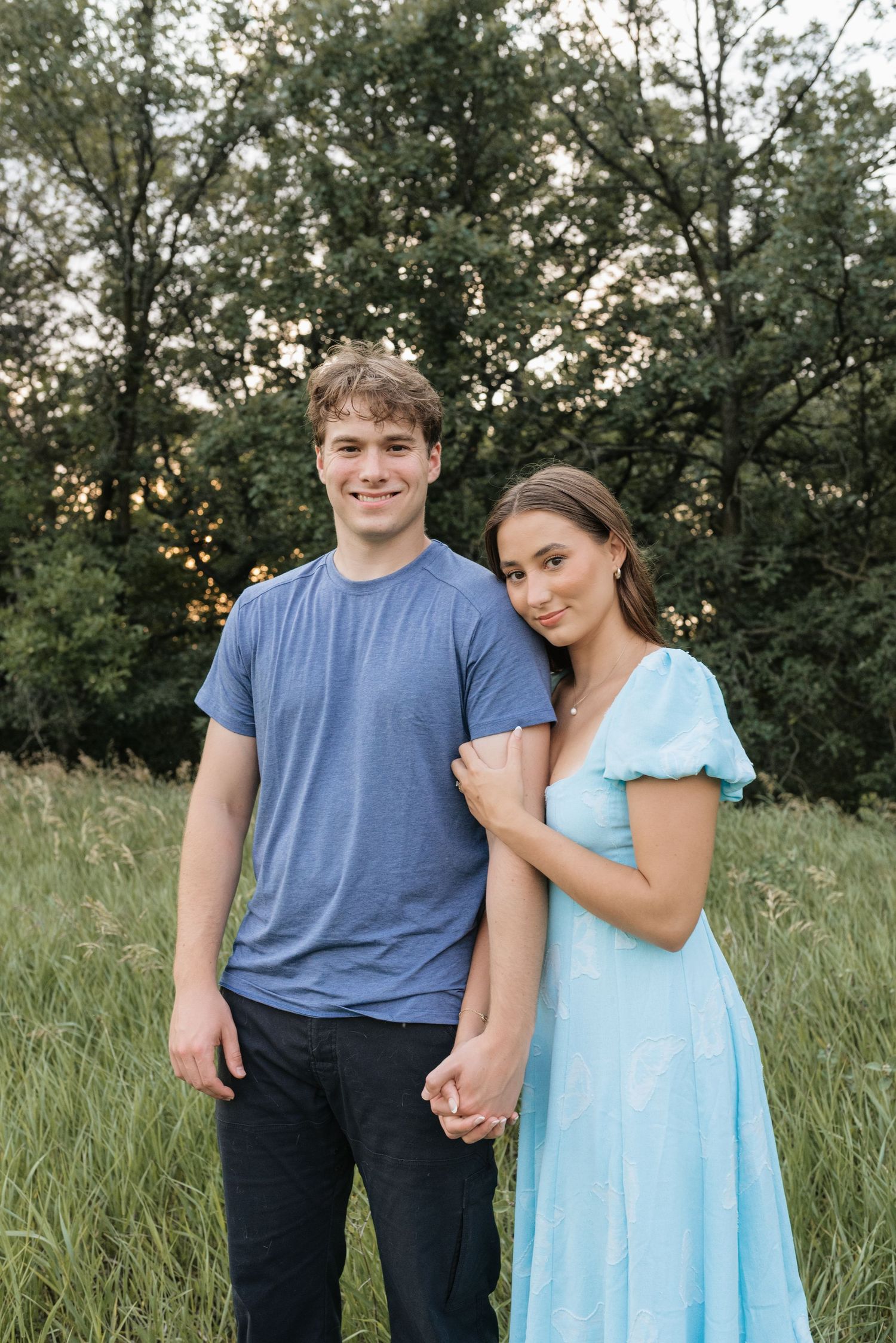 A young couple holds hands in a lush green field, smiling, wearing a blue t-shirt and light blue maxi dress.