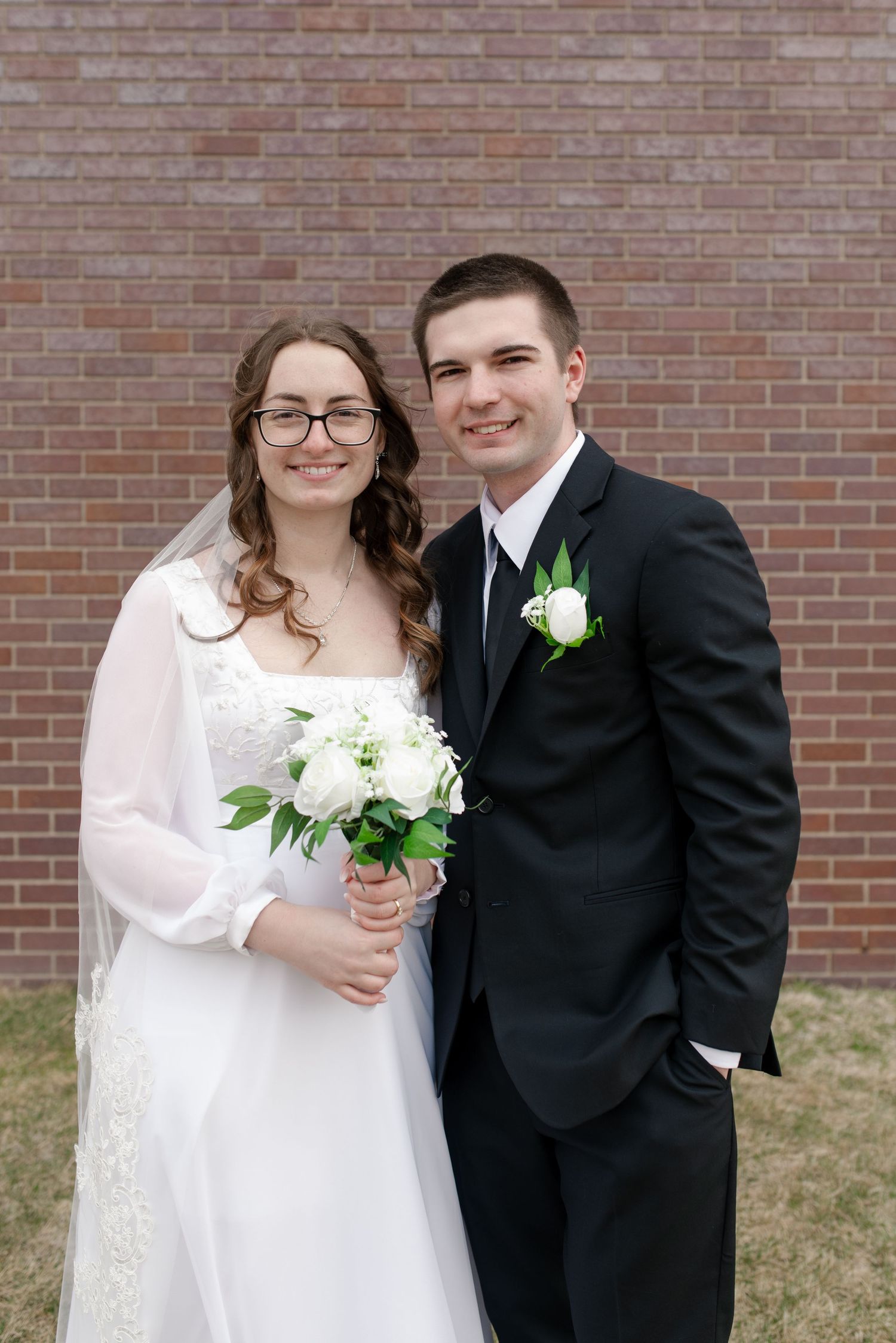 Smiling newlyweds pose before brick wall, bride holding white bouquet in lace wedding dress.