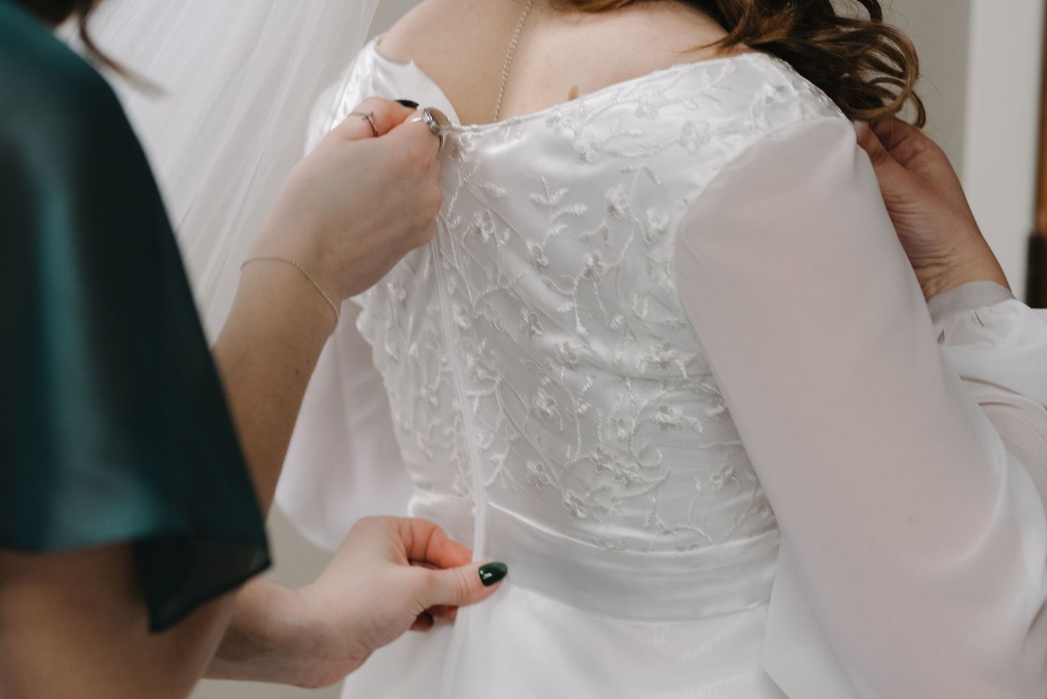 Bridesmaid fastening the back of a white lace wedding dress with long sleeves on a bride.