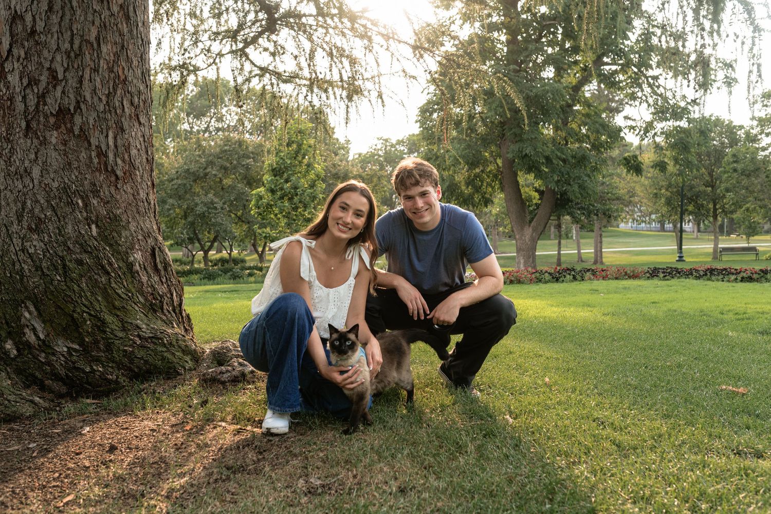 A young couple smiles with their small dog beside a tree in a lush green park during golden hour.
