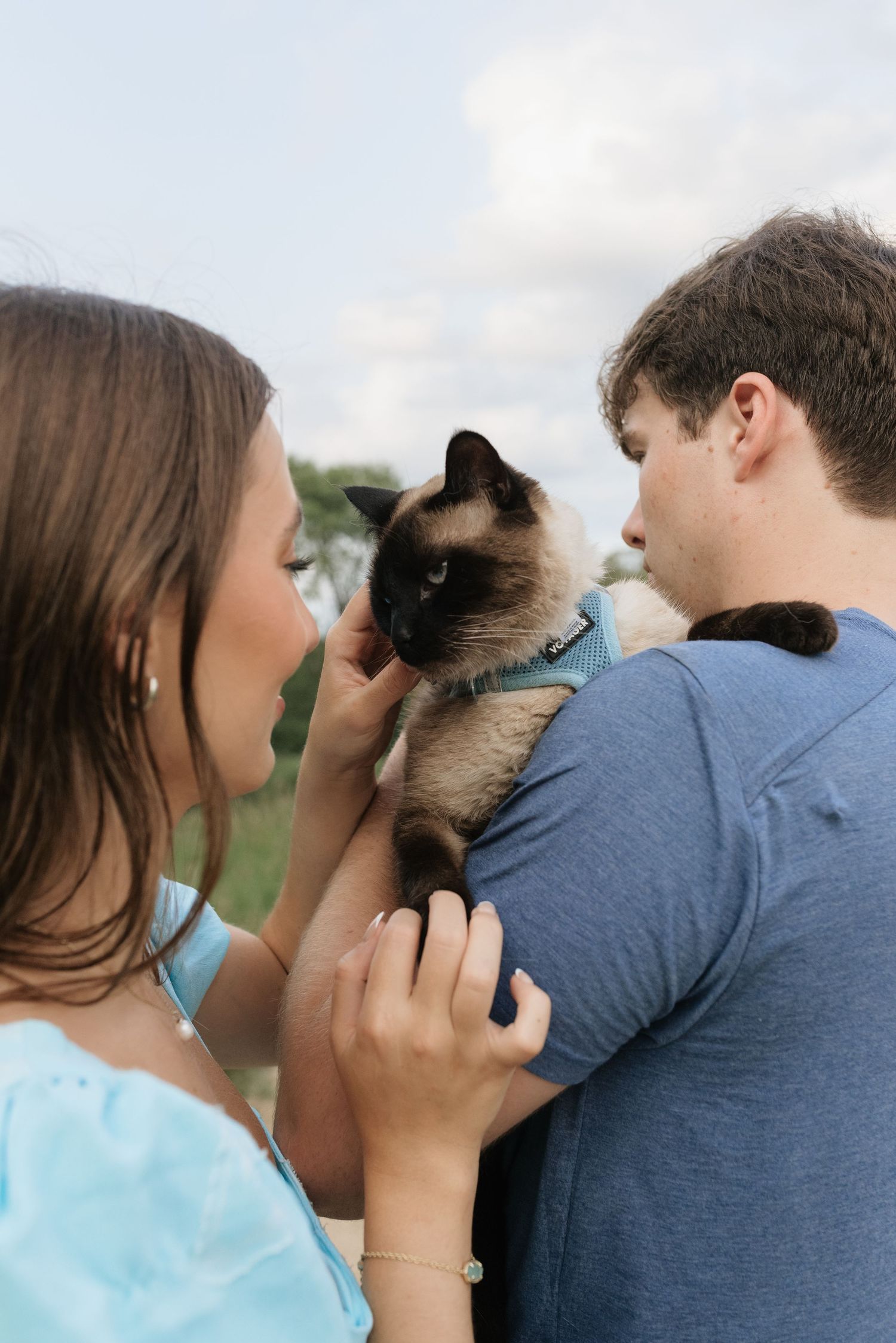 A couple holds a Siamese cat outdoors, the woman in blue gently touching the cat as the man looks on.