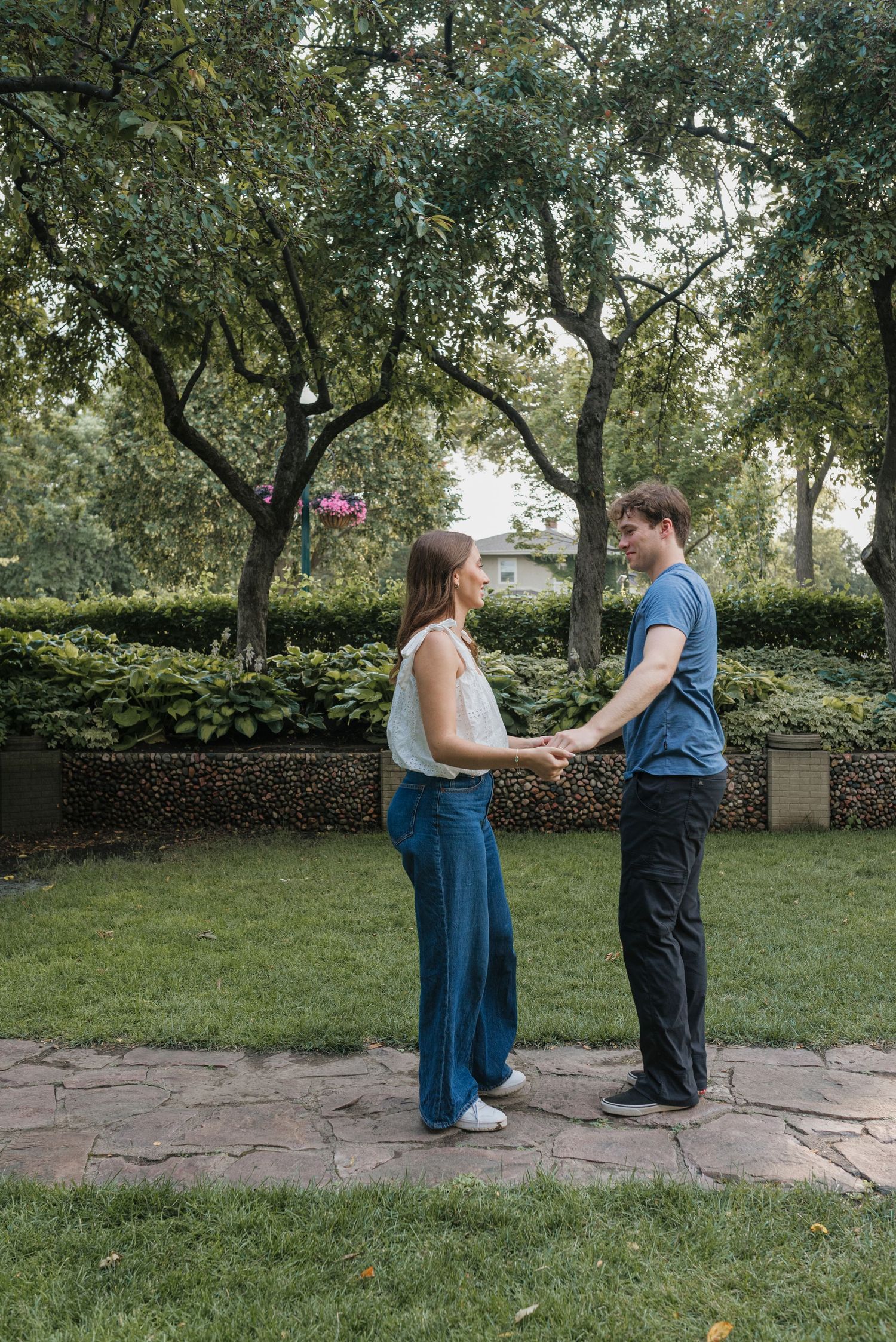 A couple holds hands facing each other on a stone path in a lush green park surrounded by trees.
