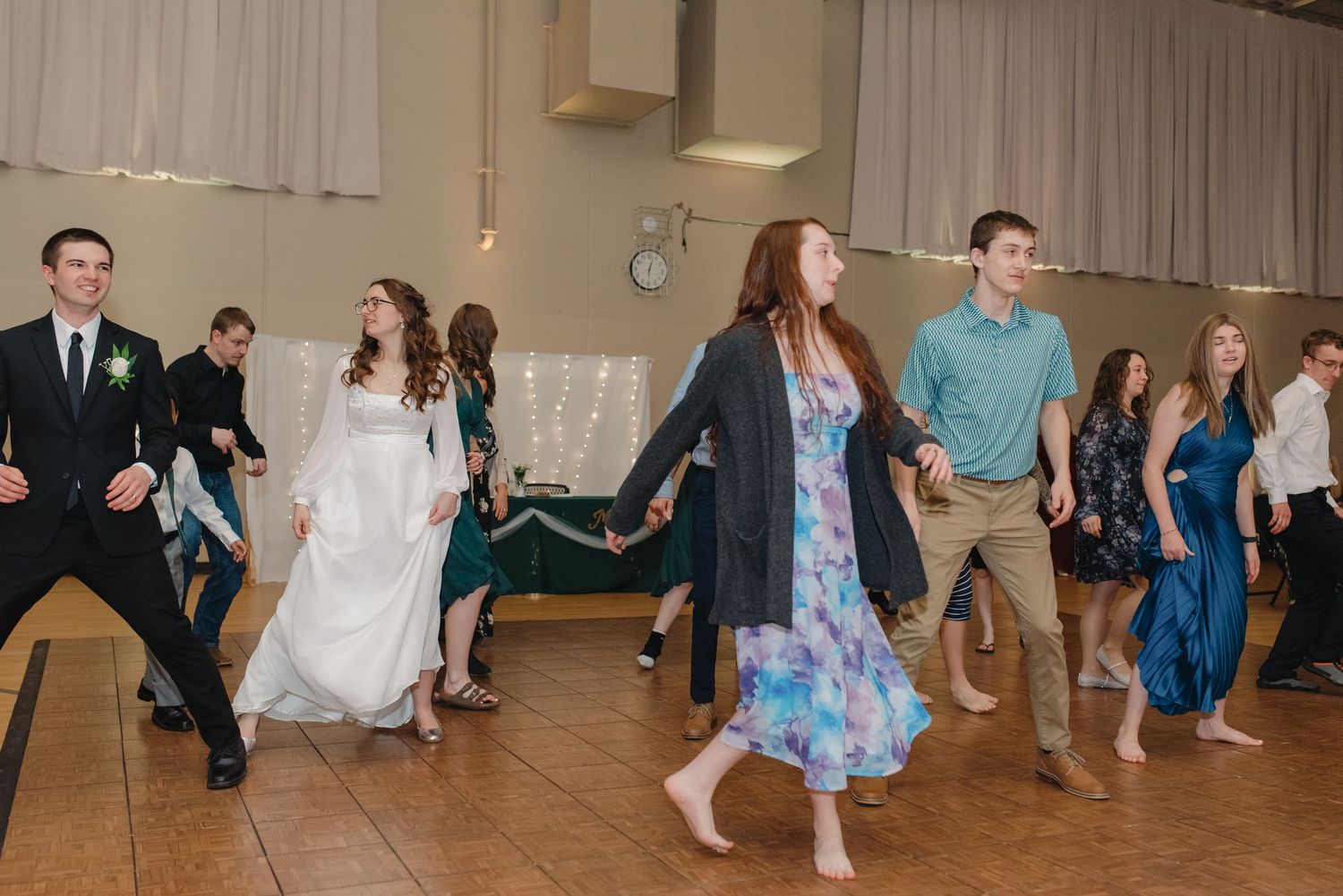 Wedding guests and bride in white dress dancing together in a line at lively reception party.