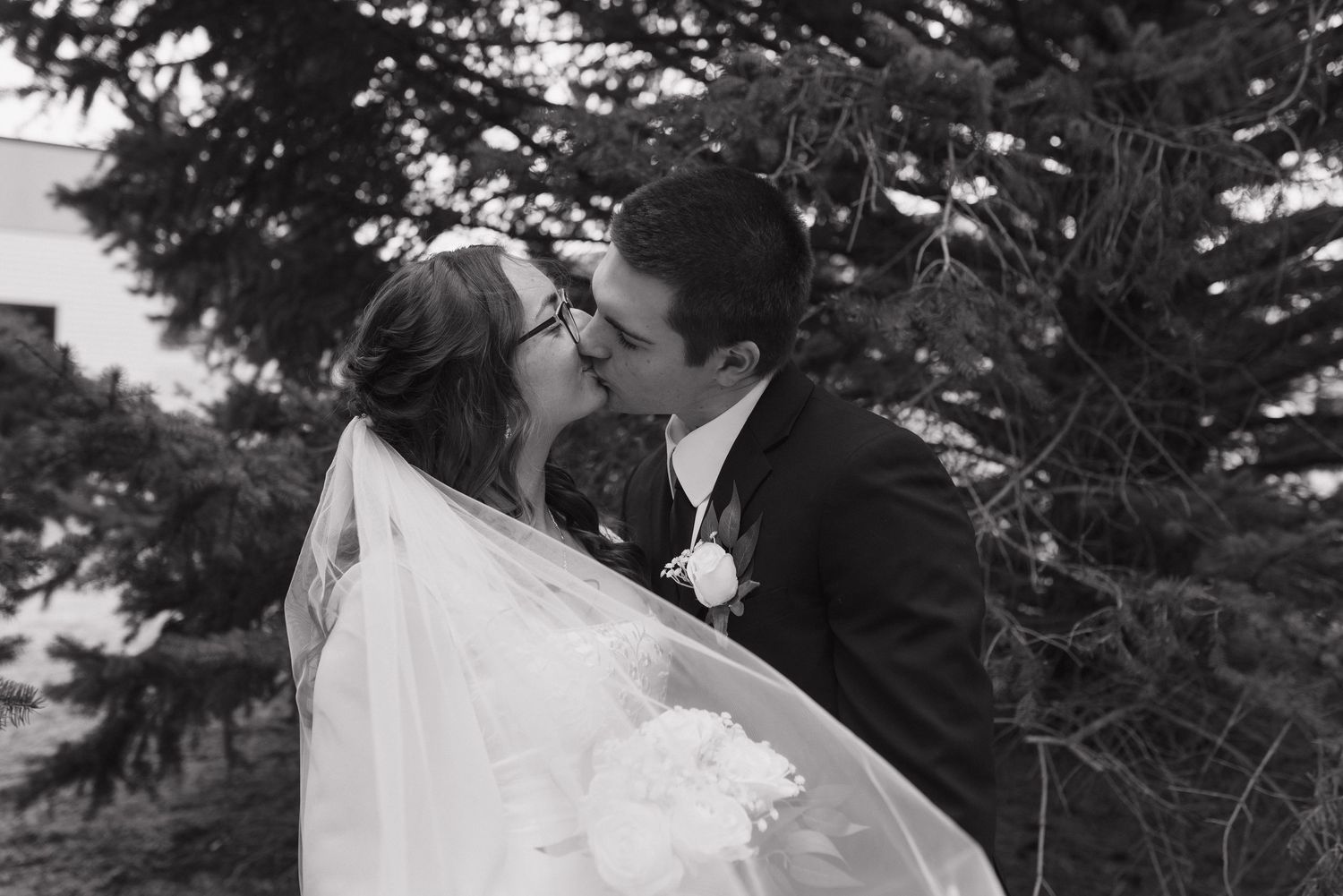 Newlyweds share a romantic kiss outdoors with the bride's veil flowing in the wind.